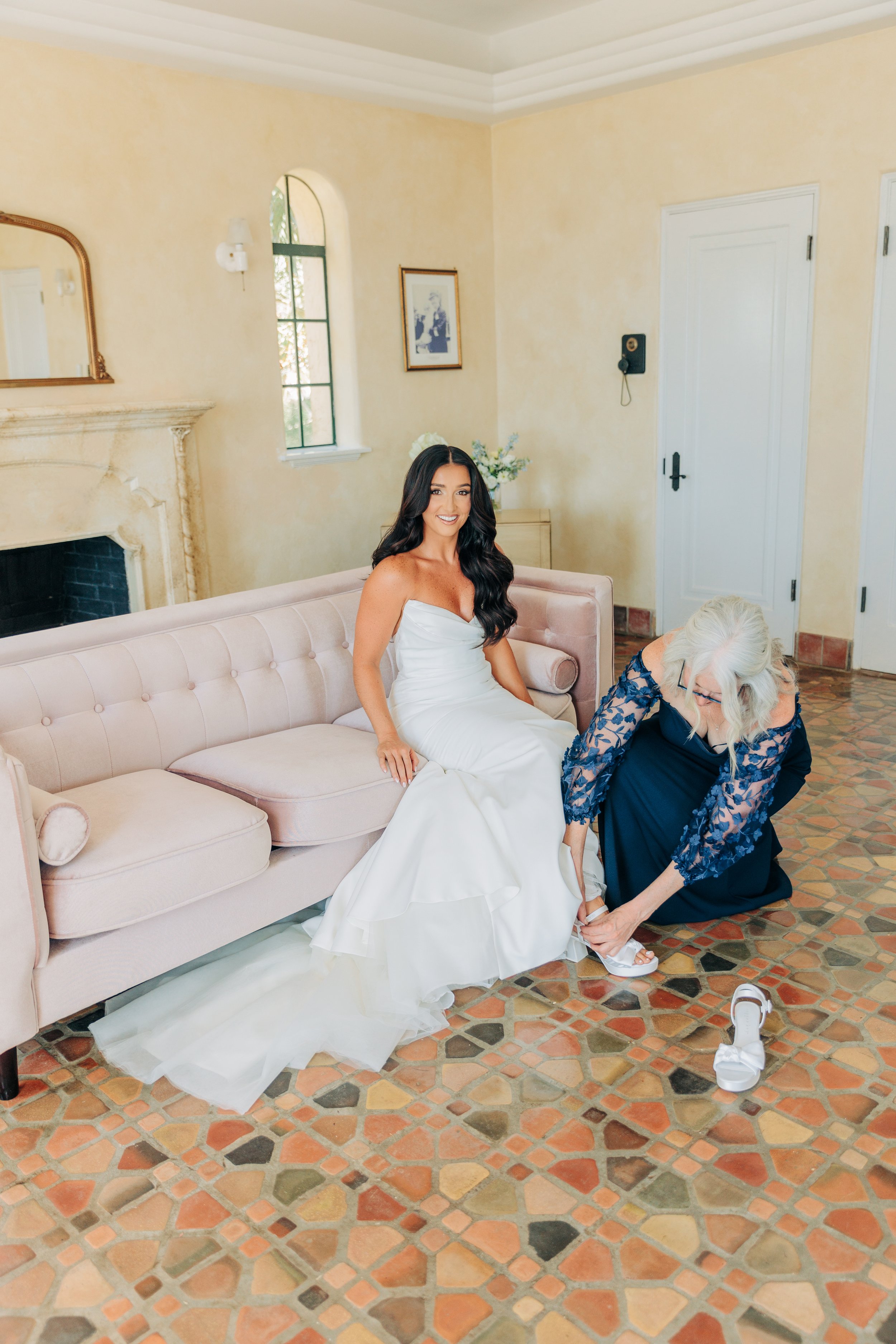 A young woman in a wedding dress smiling on a pink sofa, while an older woman adjusts her sandal in a cozy room with a stone fireplace, a window, and artwork on the wall.