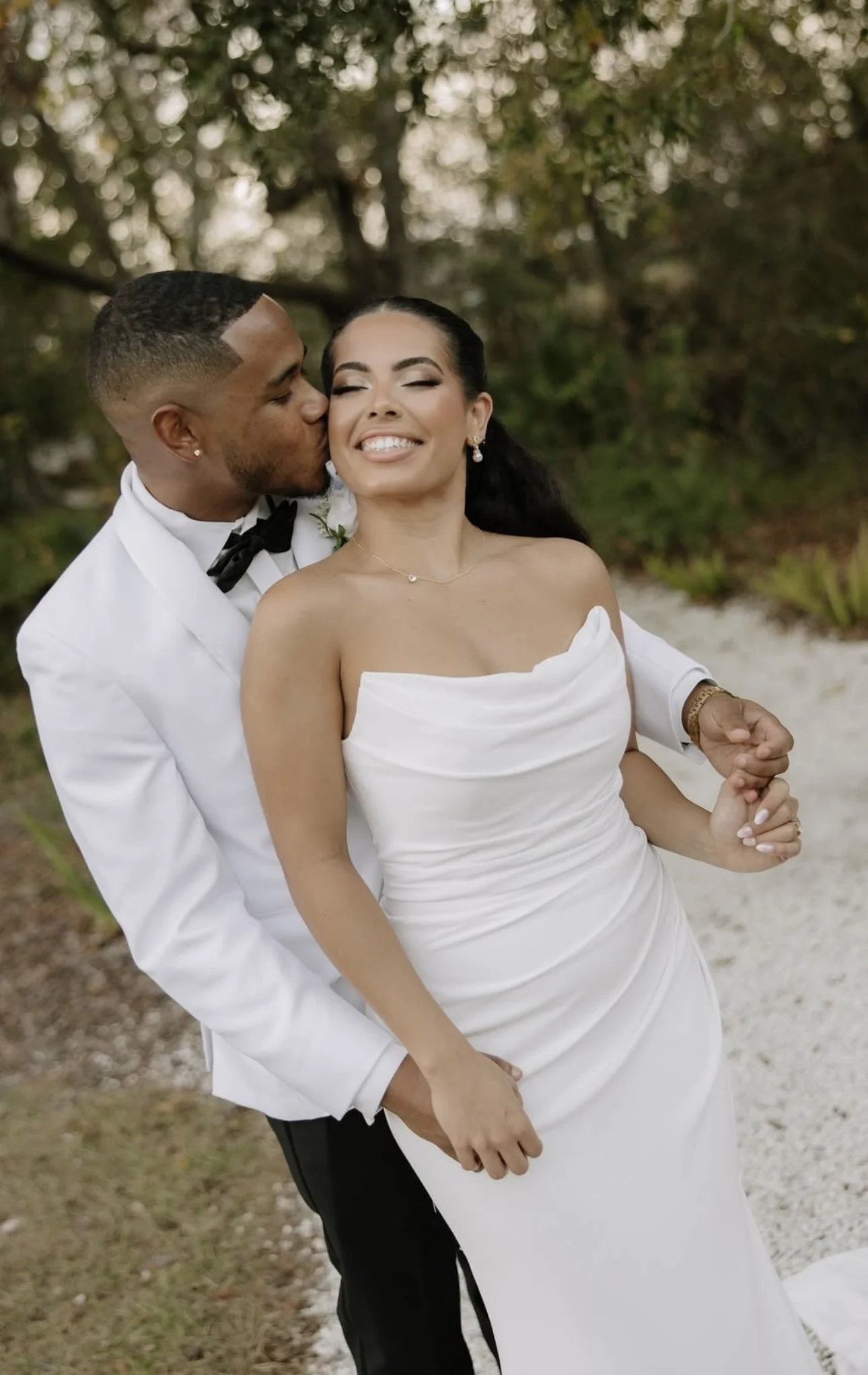 A newlywed couple outdoors, the groom kissing the bride's cheek, both smiling happily, surrounded by greenery.