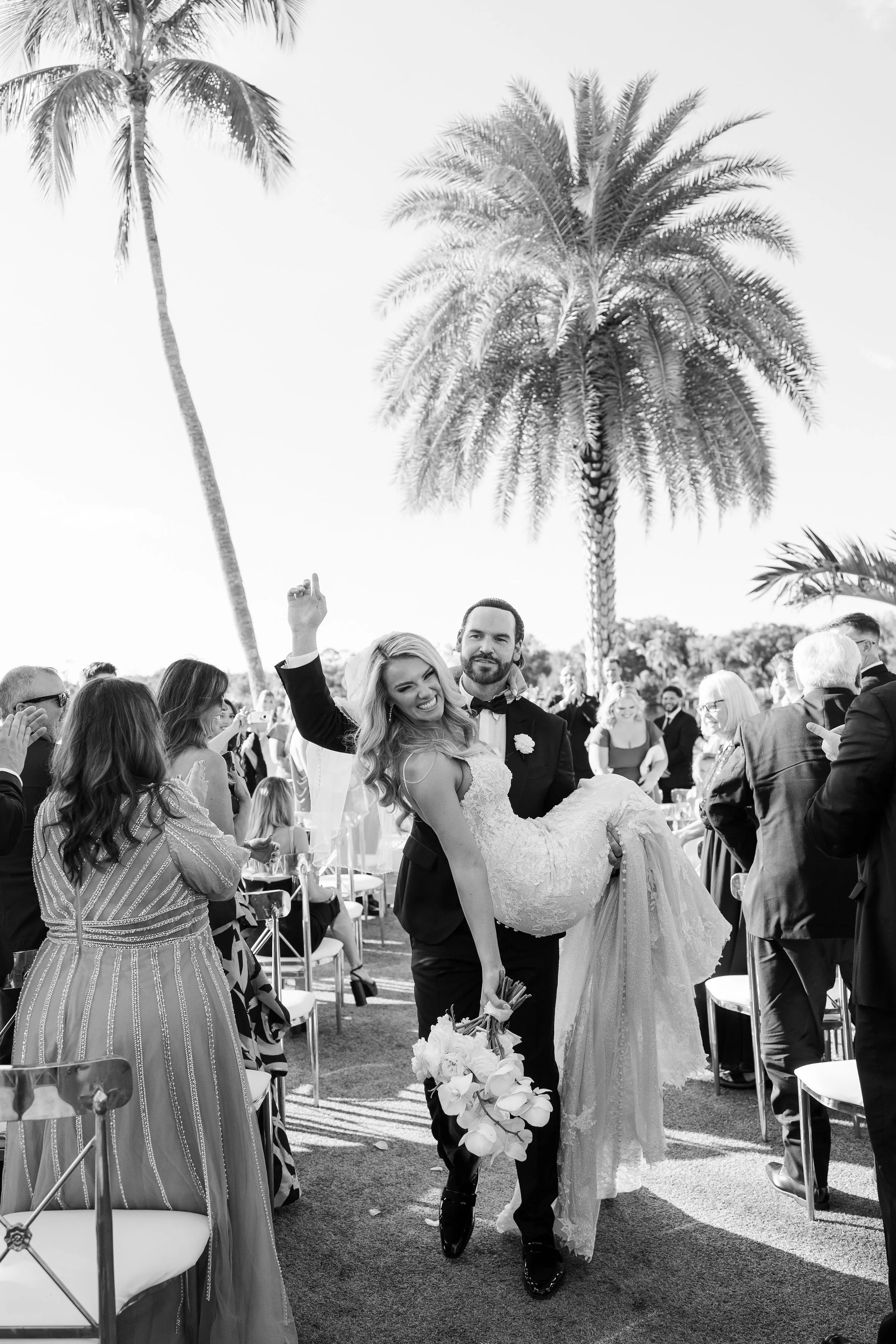 A newlywed couple with the groom carrying the bride, surrounded by wedding guests at an outdoor wedding reception with palm trees in the background.