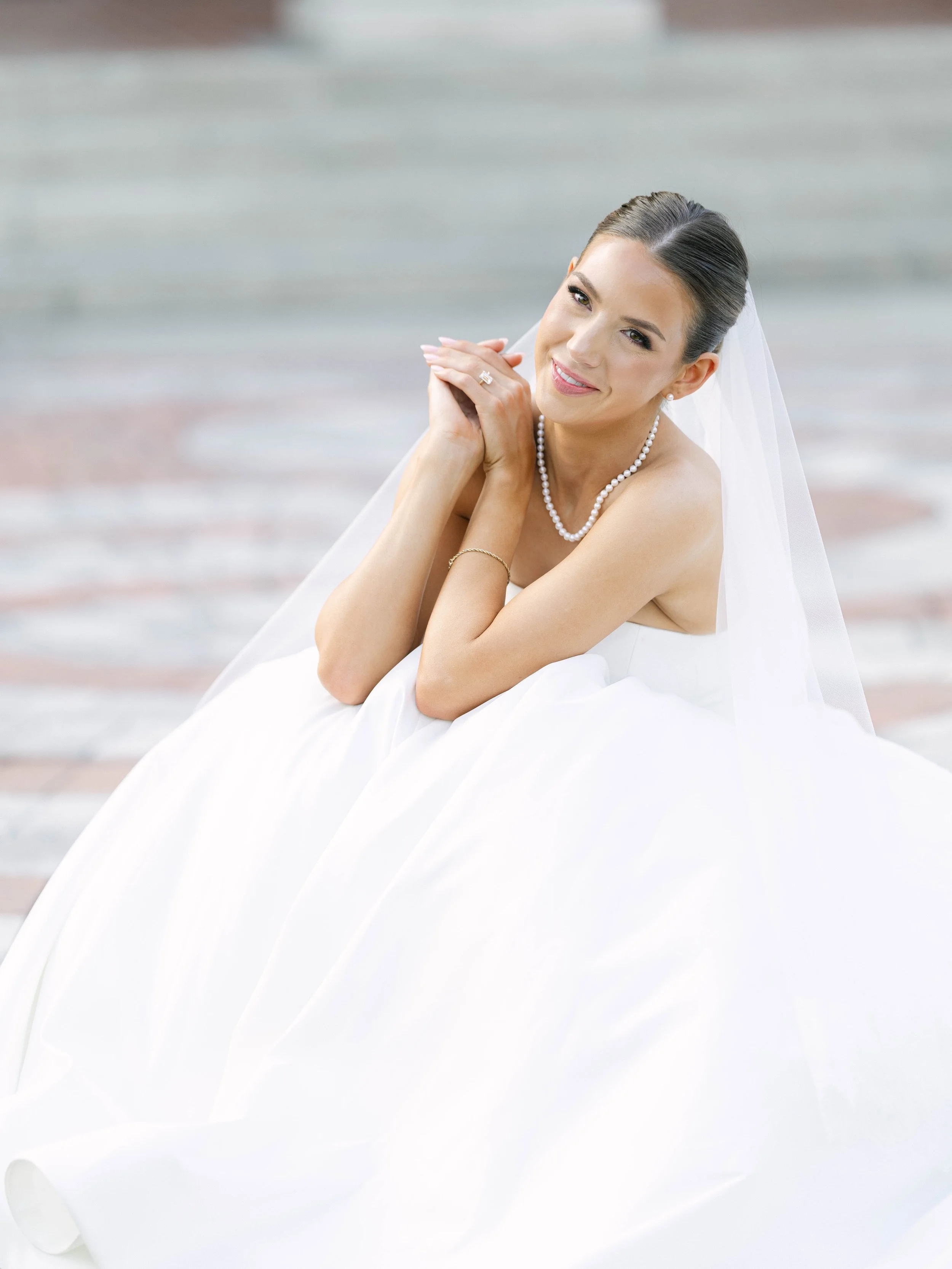 A portrait of a smiling bride with dark hair styled in an elegant updo, wearing a pearl necklace, pearl earrings, a wedding dress, and a veil, posing outdoors on a soft, neutral background.