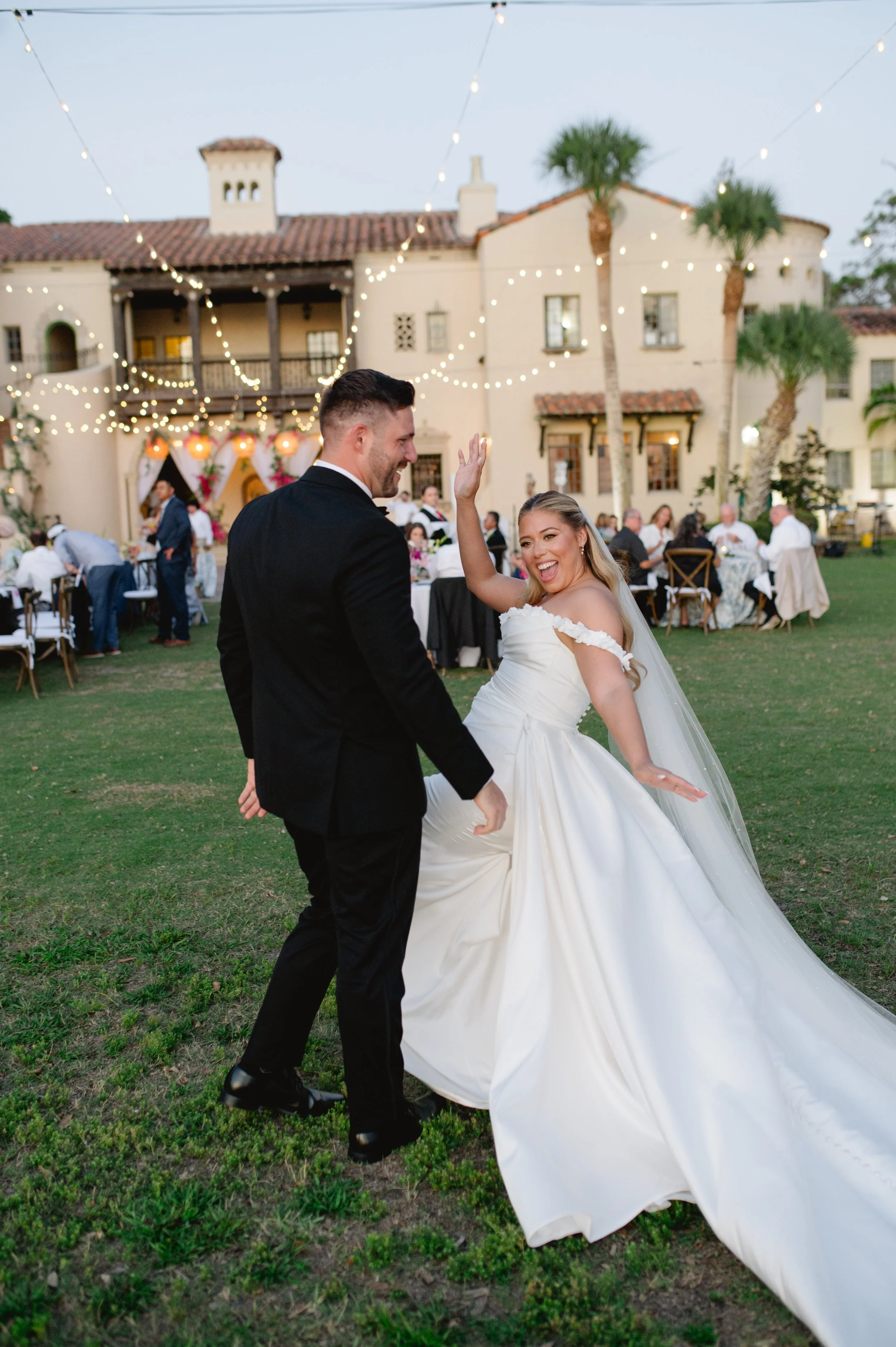 A newlywed couple dancing outdoors at their wedding reception during sunset, with guests seated at decorated tables, string lights overhead, and a large house in the background.