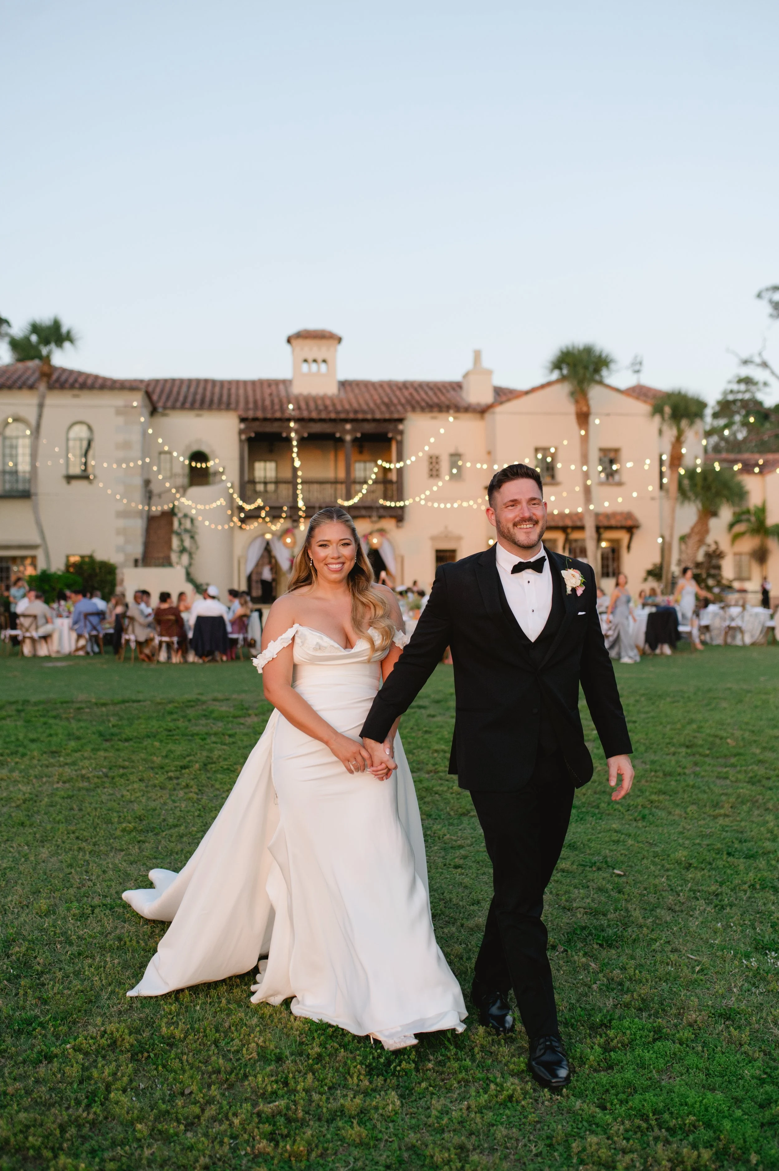 A newlywed couple walking hand in hand during their outdoor wedding reception at sunset, with string lights illuminating a Spanish-style building in the background.