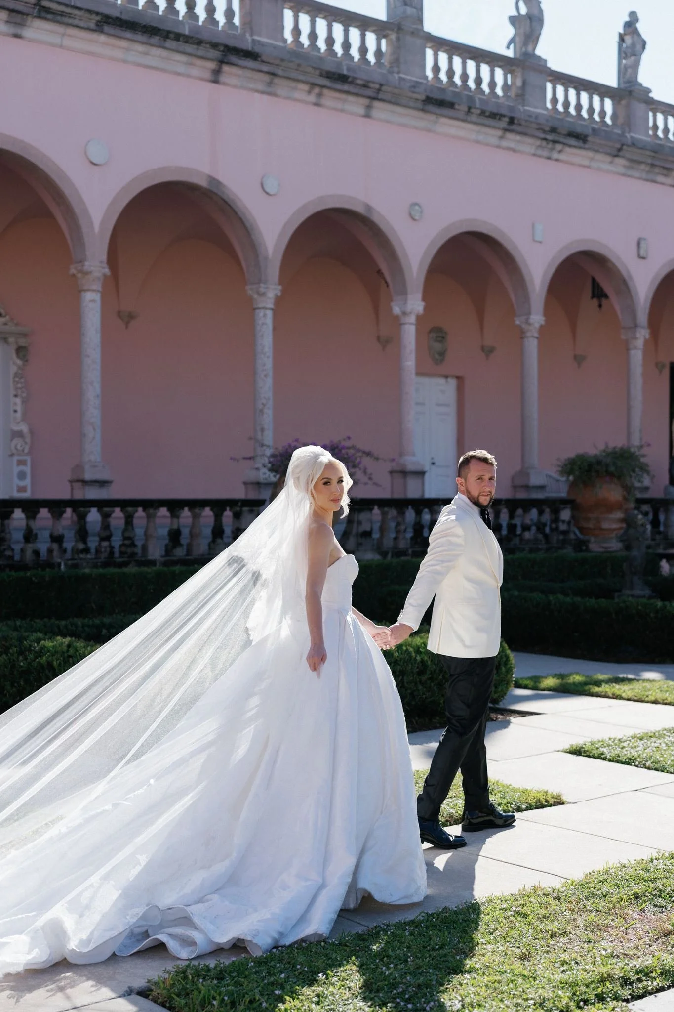A bride and groom holding hands outside in front of a pink building with arches and statues, dressed in wedding attire.