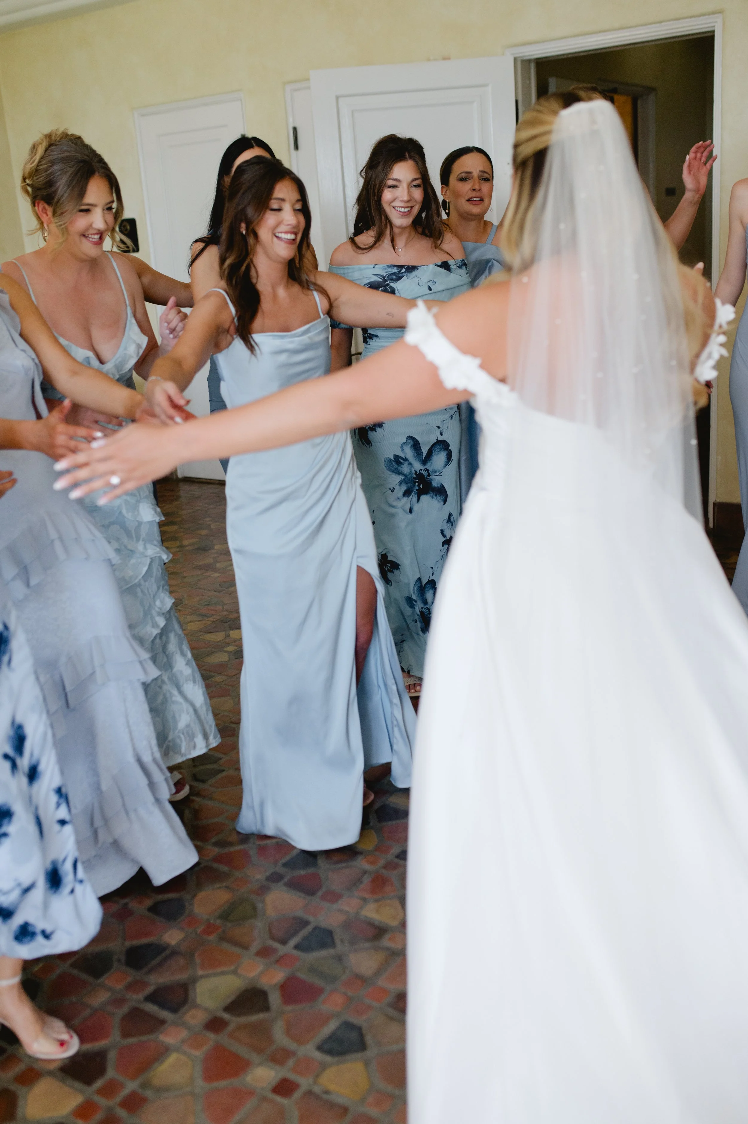 A bride with a veil and a white dress is with a group of women in blue and white dresses, dancing and celebrating in a room with a tile floor and white doors.