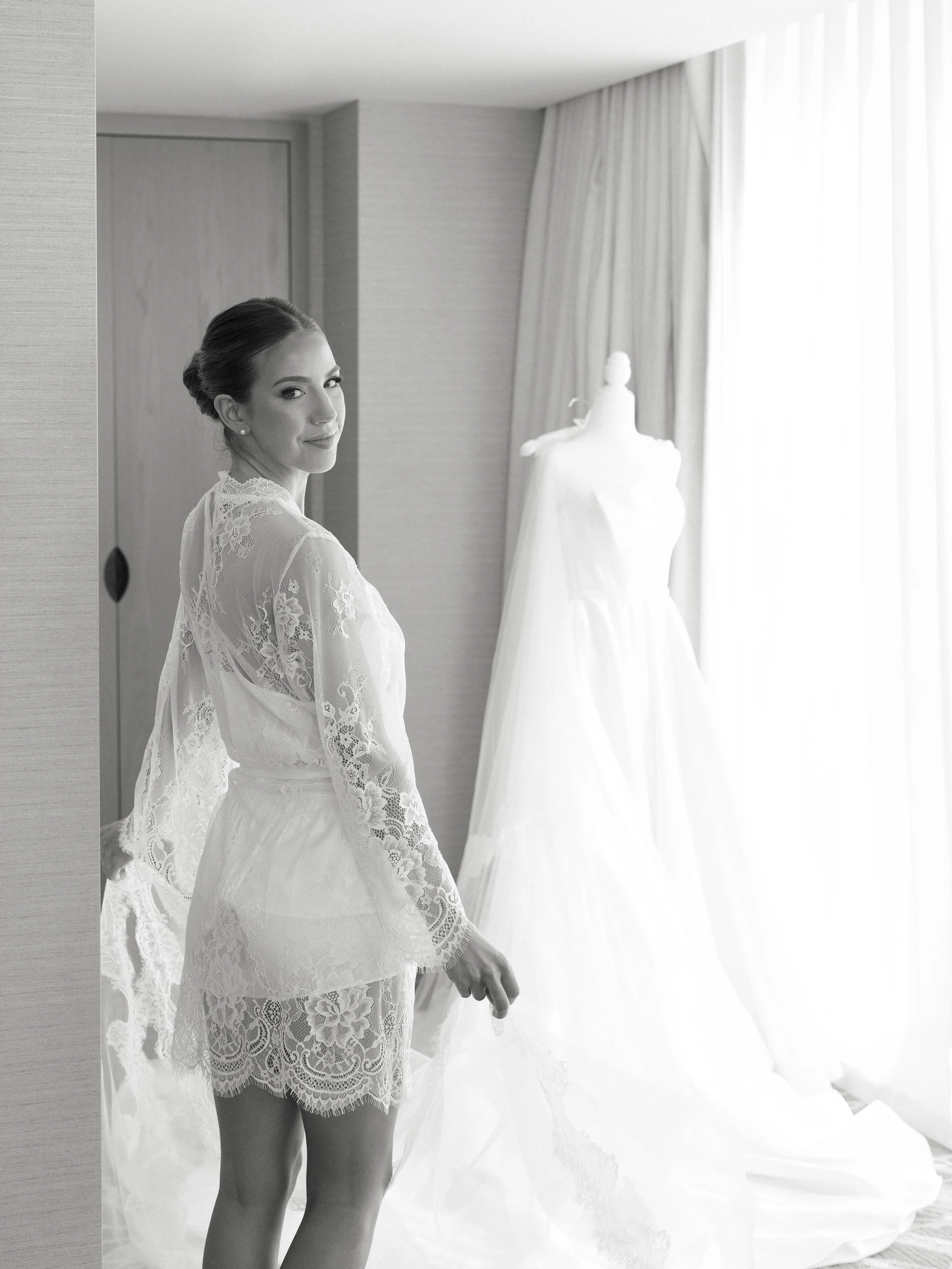 A woman in lace bridal lingerie standing in a hotel room, with a wedding dress on a mannequin beside her and sunlight streaming through curtains.