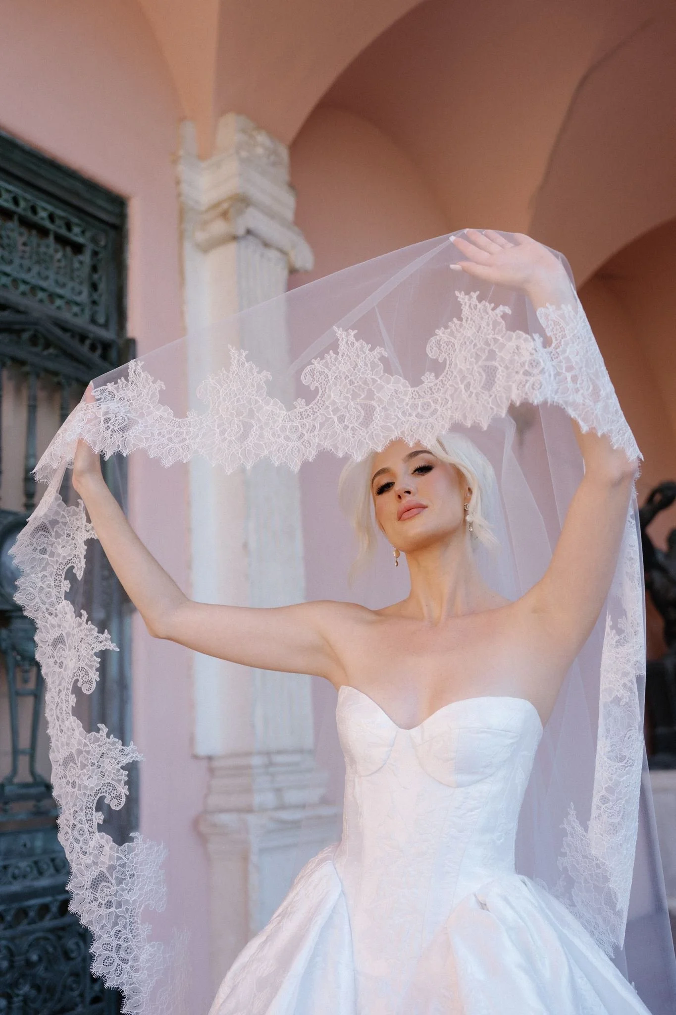A woman in a white wedding dress holding up a lace wedding veil in front of a pink building with architectural details.