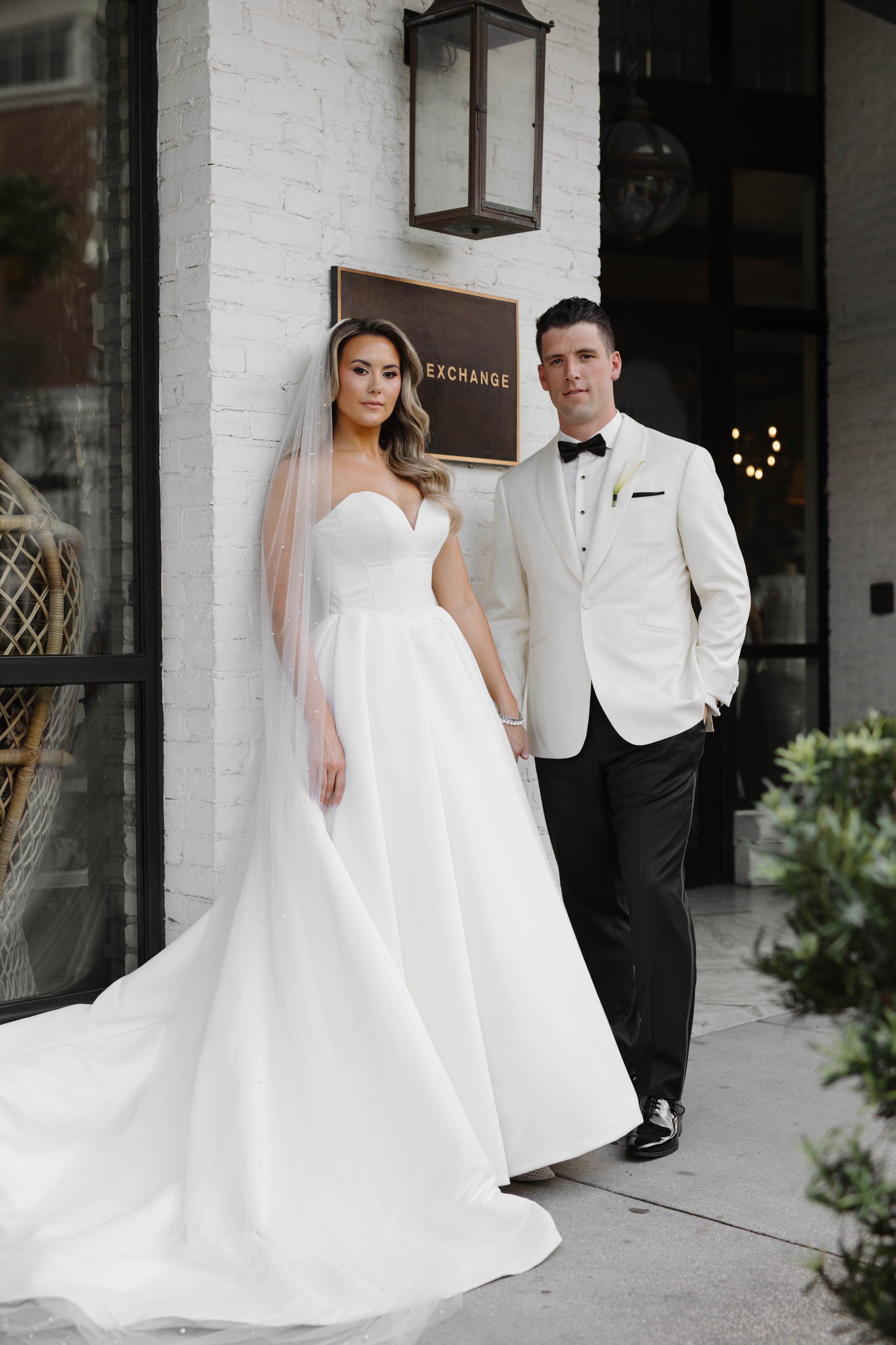 Bride in a white wedding dress with a veil and groom in a tuxedo with a black bow tie standing outside a building with a sign that says 'Exchange'.