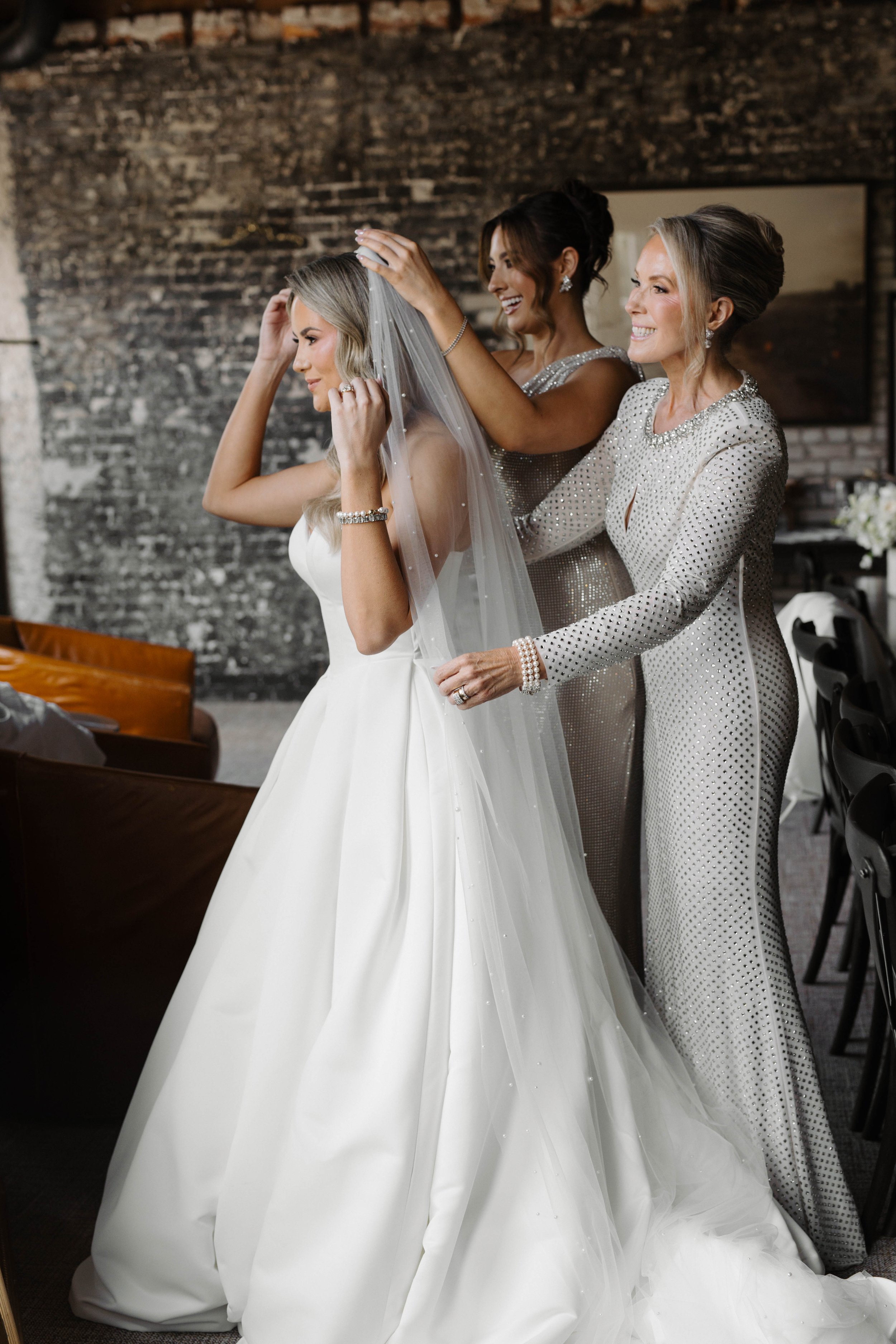 A bride wearing a white wedding gown and veil, assisted by two women in elegant, sparkling dresses, preparing for her wedding in an indoor setting with a brick wall in the background. at Oxford exchange in Tampa fl . 
