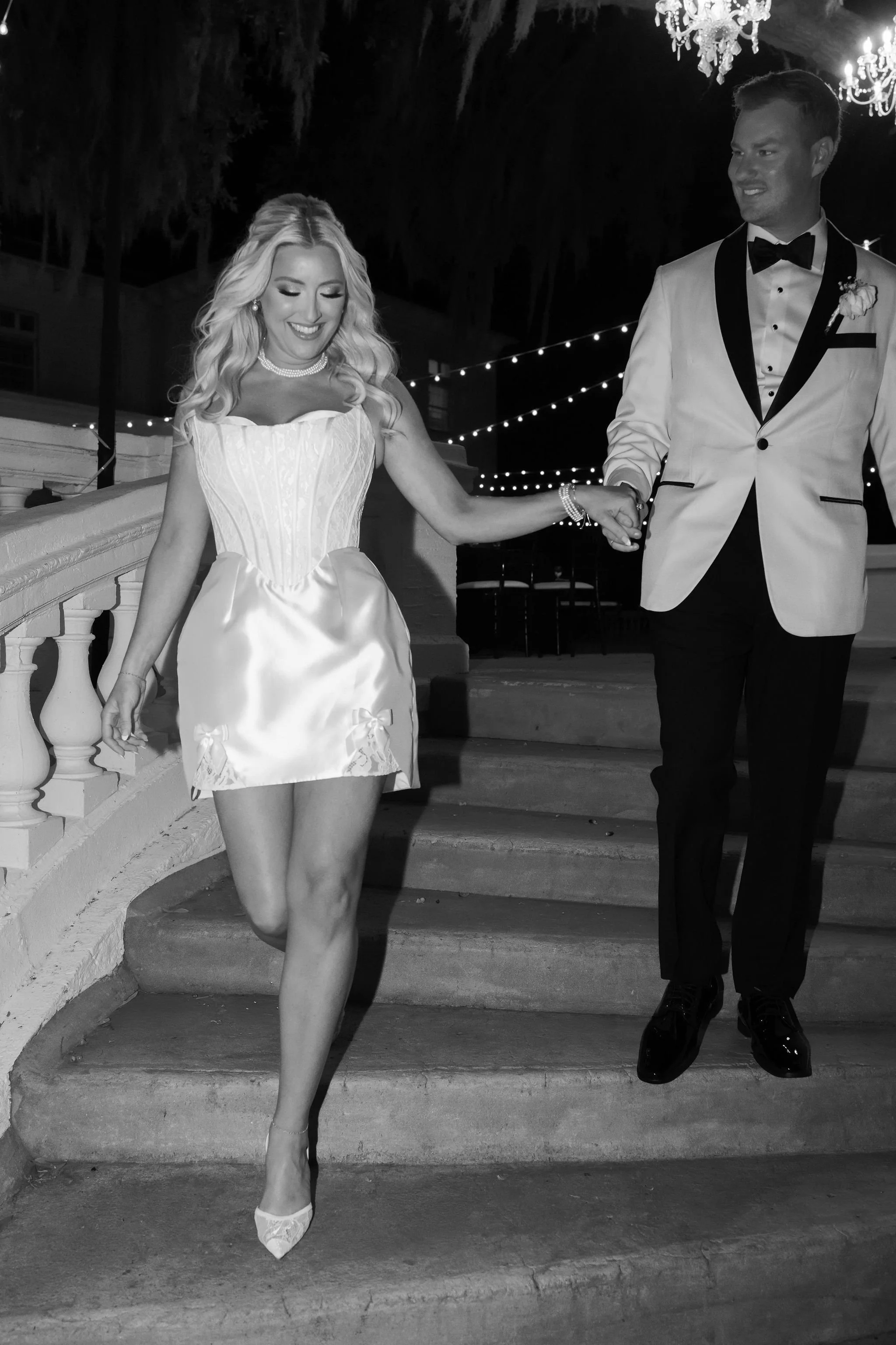 A black and white photo of a bride and groom walking down stairs at night, holding hands. The bride is smiling and wearing a strapless wedding dress and high heels, while the groom is looking at her and wearing a tuxedo with a bow tie.