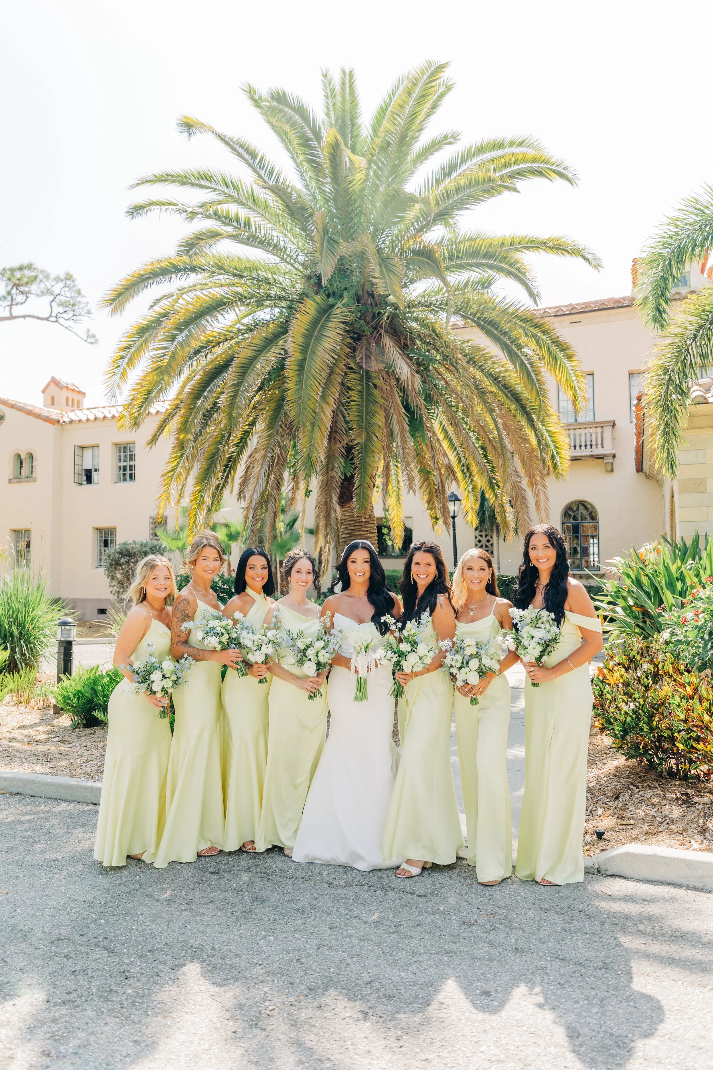 Group of eight women in matching light yellow dresses posing outdoors in front of a large palm tree, with a building in the background.