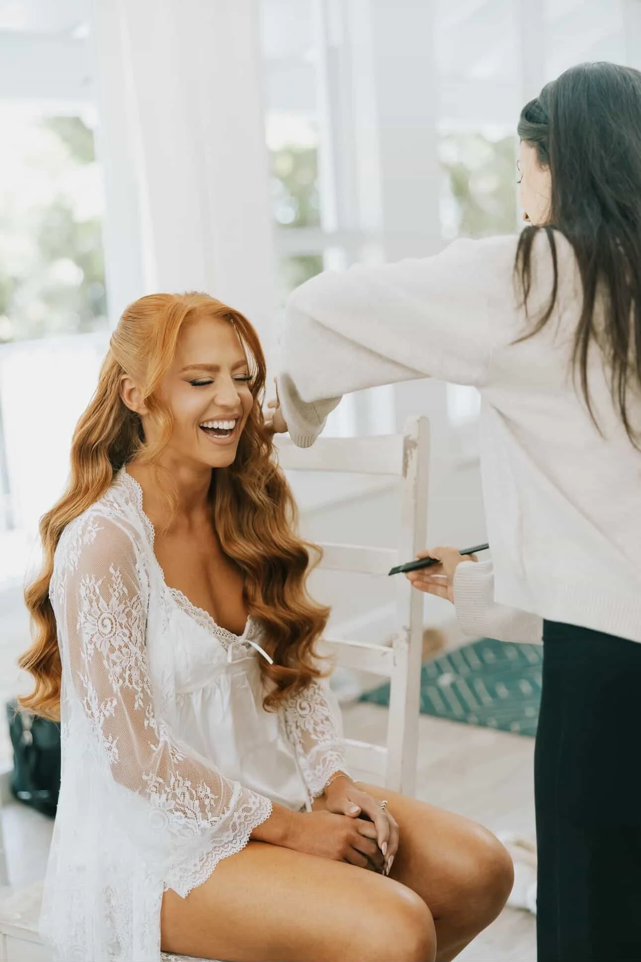 A woman with long, wavy red hair and a white lace dress is sitting and smiling while another woman with dark hair and glasses is applying makeup or taking a photo of her in a bright, airy room.