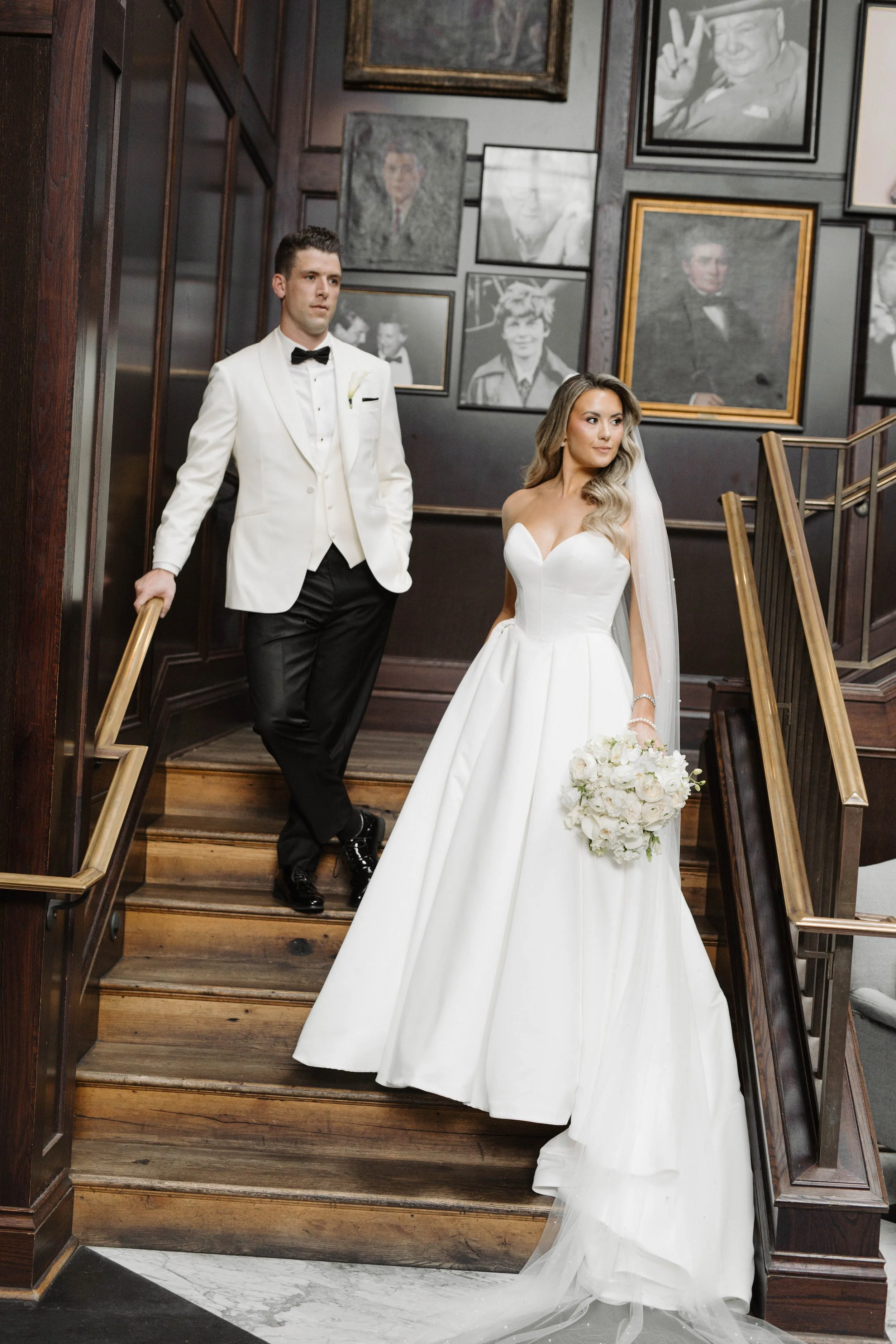 A bride in a white wedding dress holding a bouquet of white flowers stands on a wooden staircase, while a groom in a white tuxedo with a black bow tie stands behind her, holding the staircase railing. The background features framed black-and-white ph
