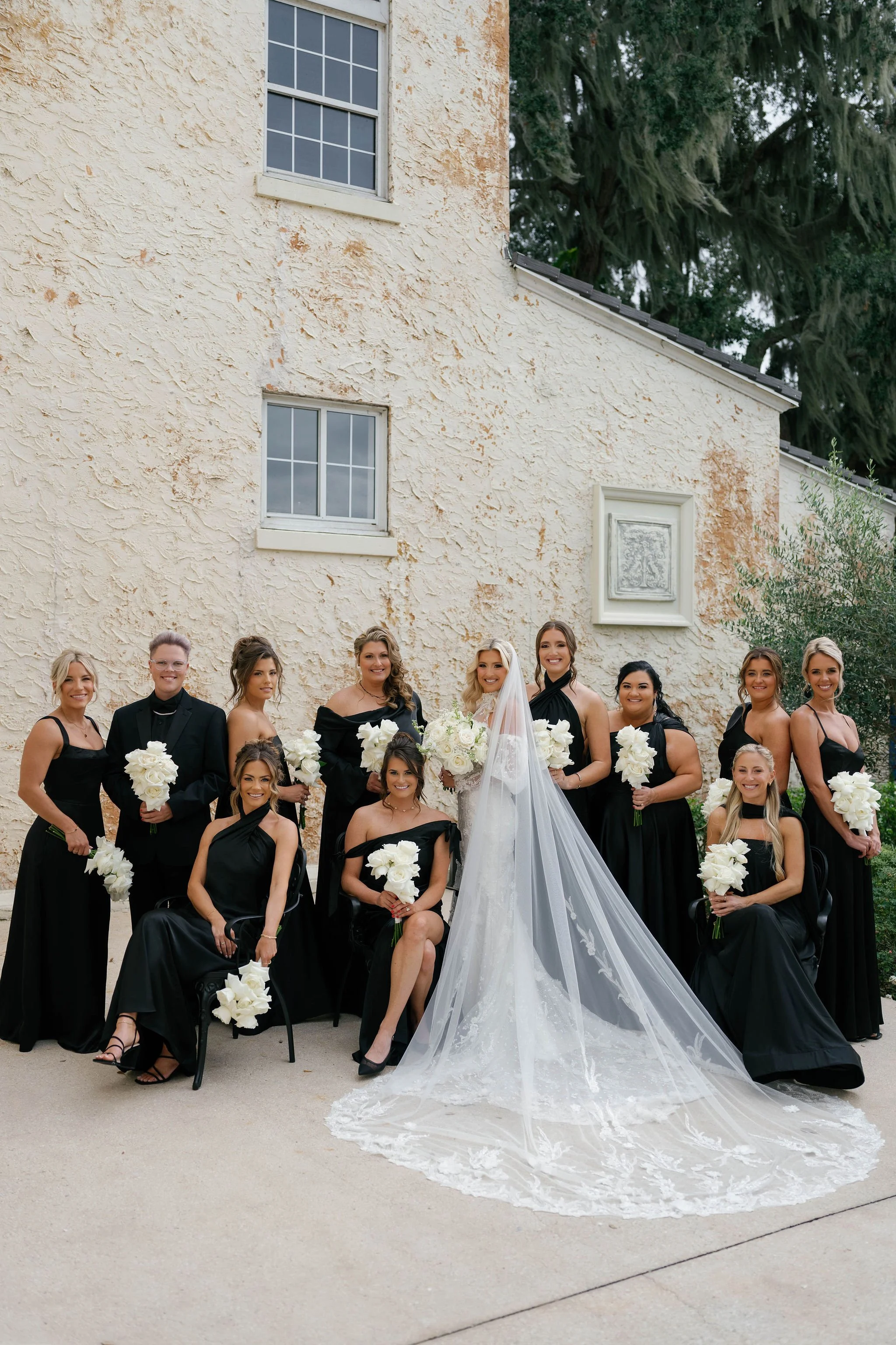 A group of women and a bride dressed in black gowns, posing outdoors in front of a beige stucco building with windows, holding white flowers with green leaves, with a large tree in the background.