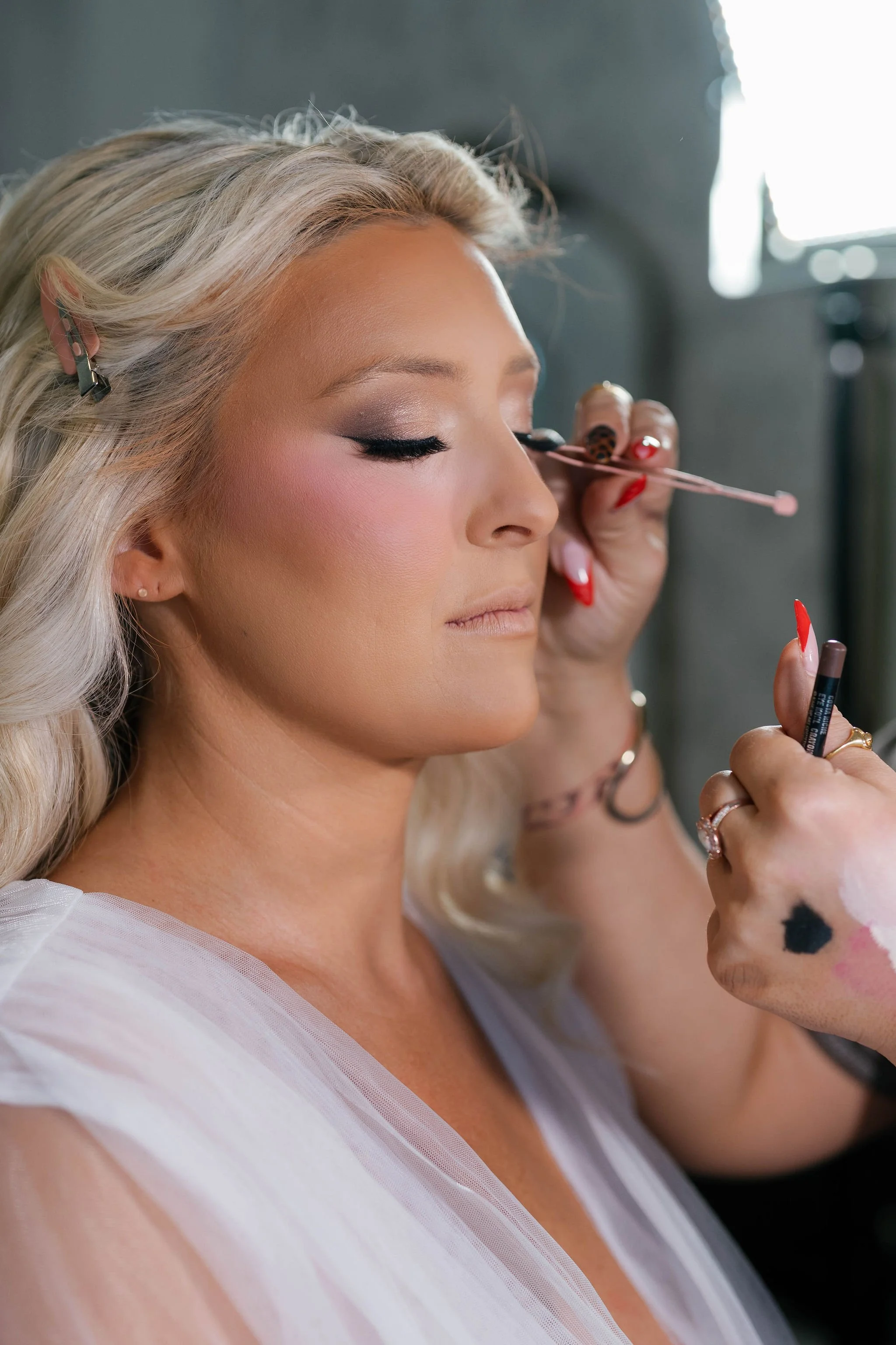 A woman with blonde hair getting her makeup done, with her eyes closed, in a professional setting.