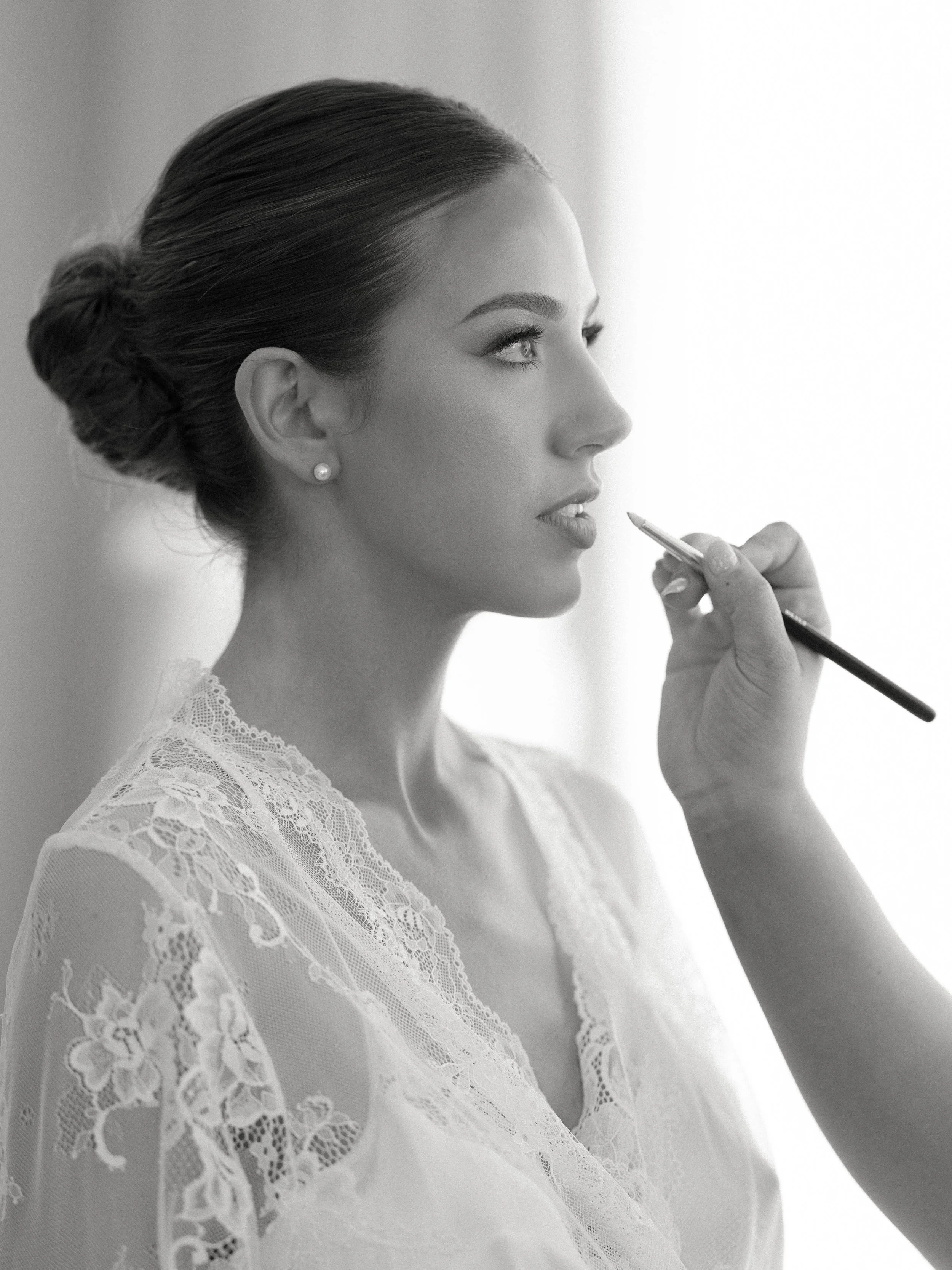 Black and white photo of a woman in profile with her hair in a bun, wearing pearl earrings, a lace garment, and holding a makeup brush near her lips.