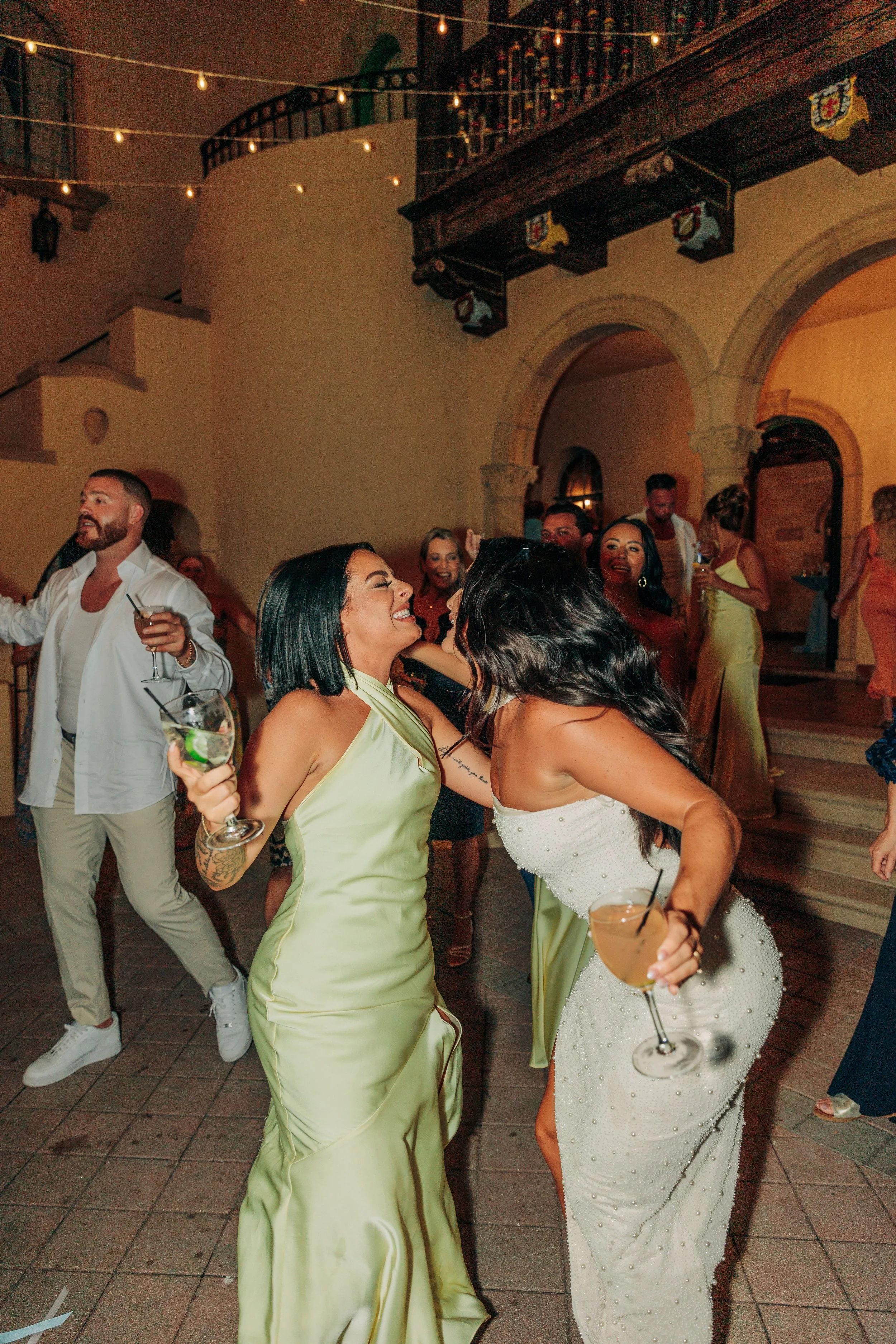 People dancing and celebrating at a party or wedding reception in a warm, decorated indoor setting with string lights, arched doorways, and a staircase, holding drinks and enjoying themselves.
