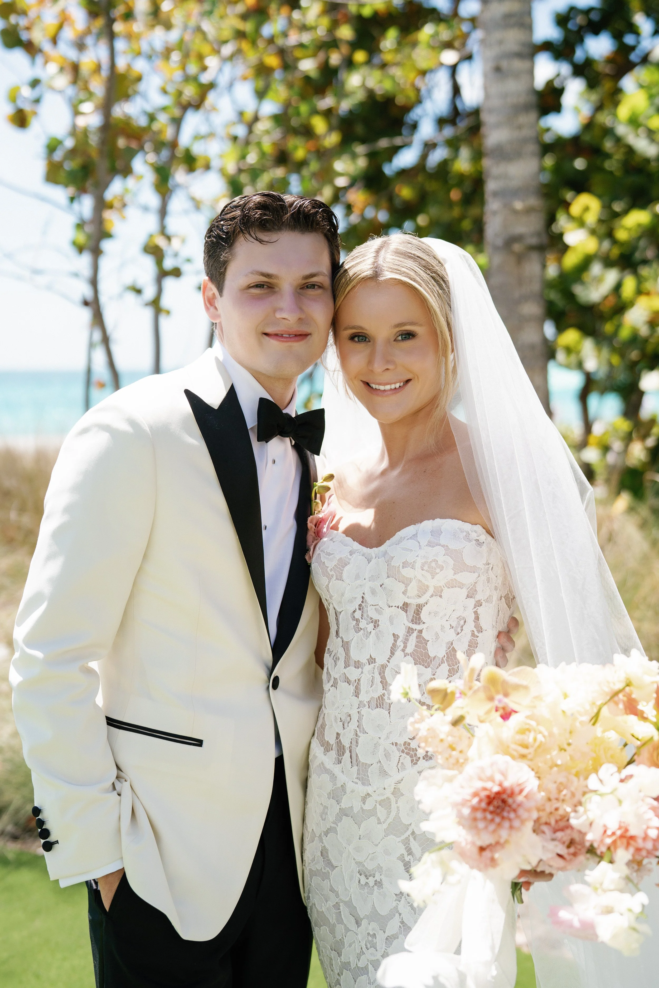 Bride and groom standing outdoors on a sunny day, smiling, with trees and a body of water in the background. The groom is wearing a white tuxedo with black accents and a black bow tie. The bride is wearing a white lace wedding dress with a veil and h