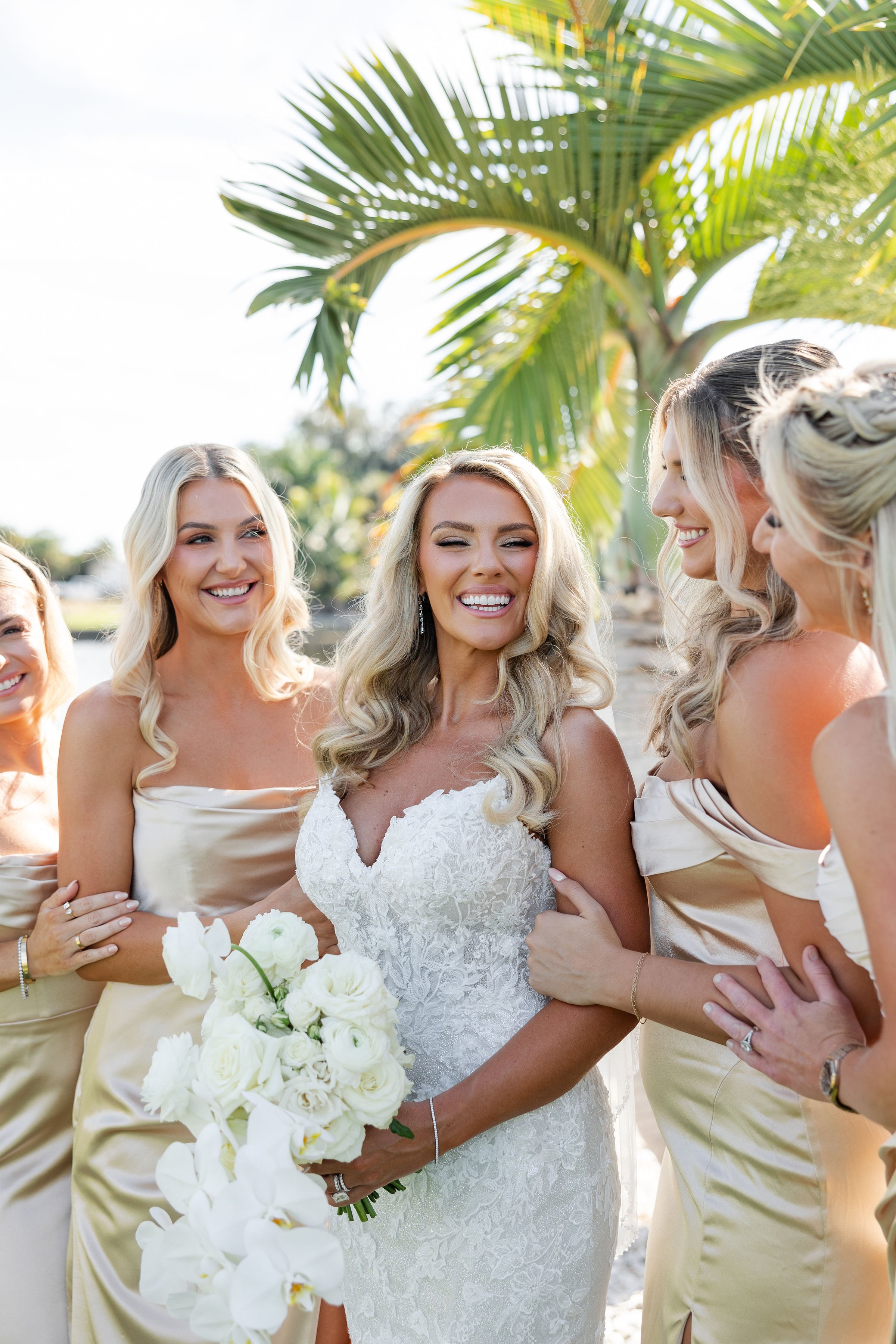 A group of women, including a bride in a white lace wedding gown holding a bouquet of white flowers, smiling and talking outdoors under a palm tree.