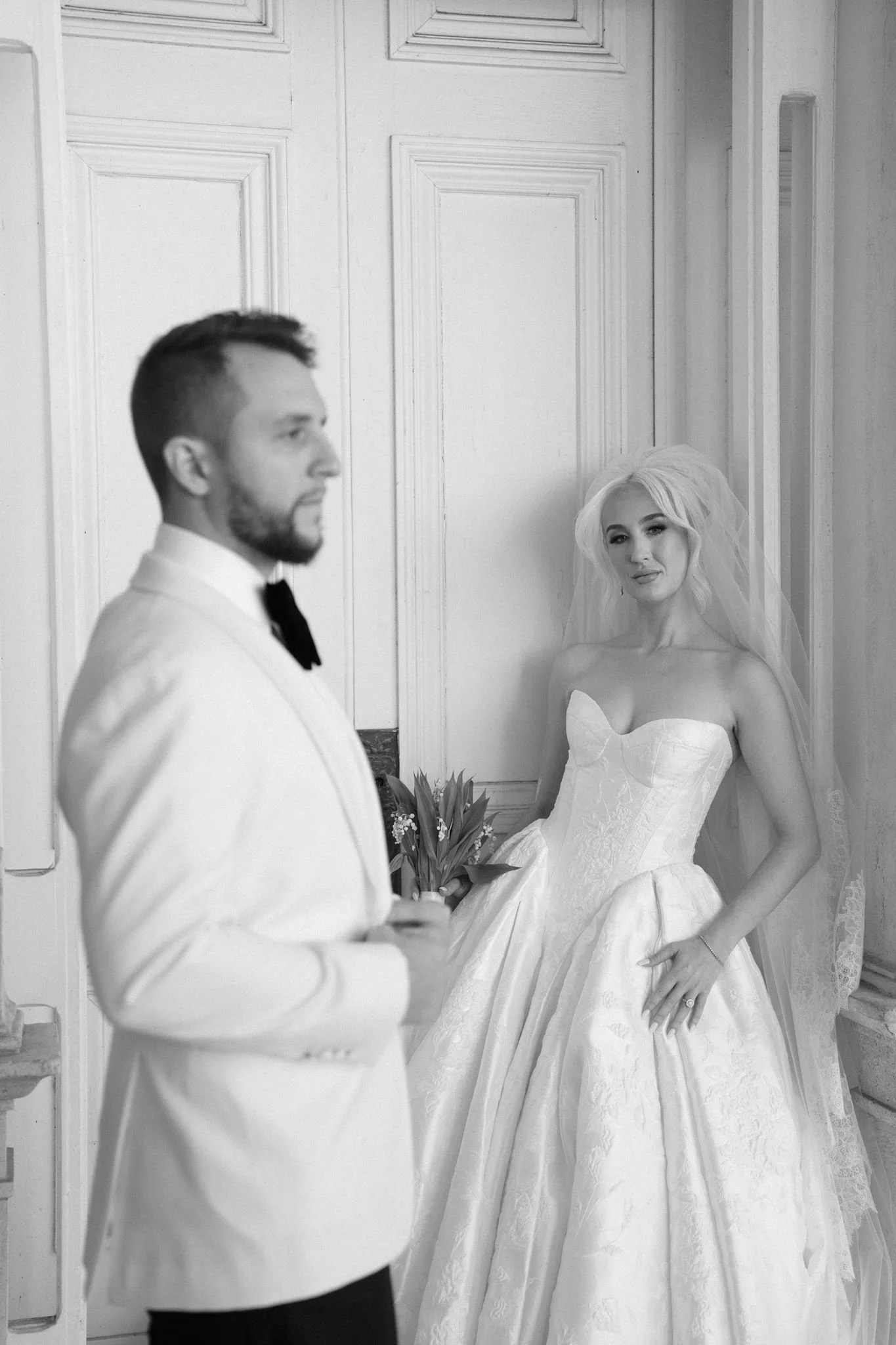A bride and groom on their wedding day indoors; the groom is in a white tuxedo with a black bowtie, looking away, holding a bouquet, while the bride, in a strapless wedding gown with a full skirt and veil, is smiling softly at the camera.