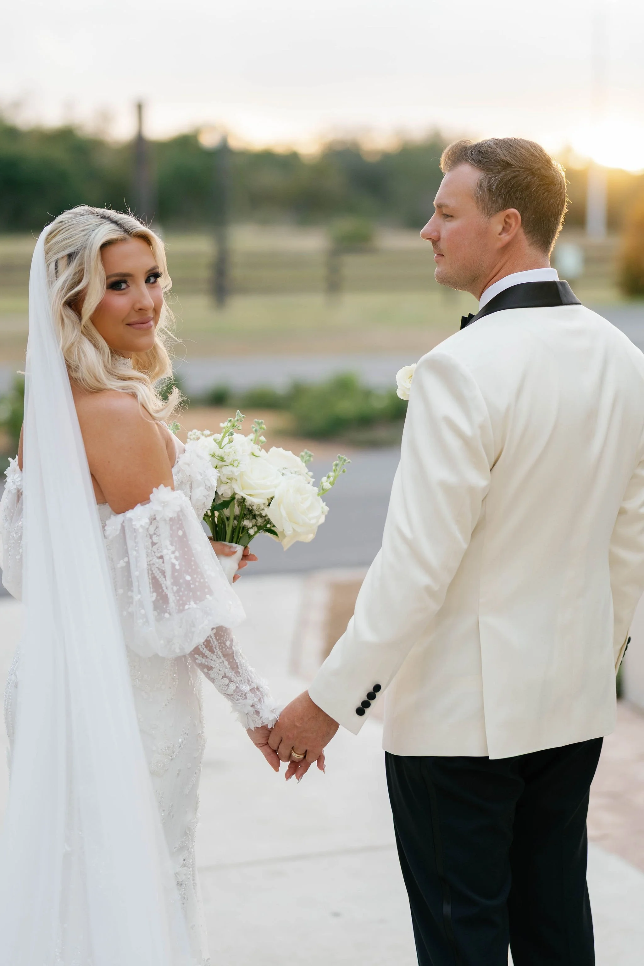 A bride and groom holding hands during their wedding ceremony outside, with the bride in a white lace dress and veil, holding a bouquet of white flowers, and the groom in a white tuxedo jacket with black pants, facing each other.