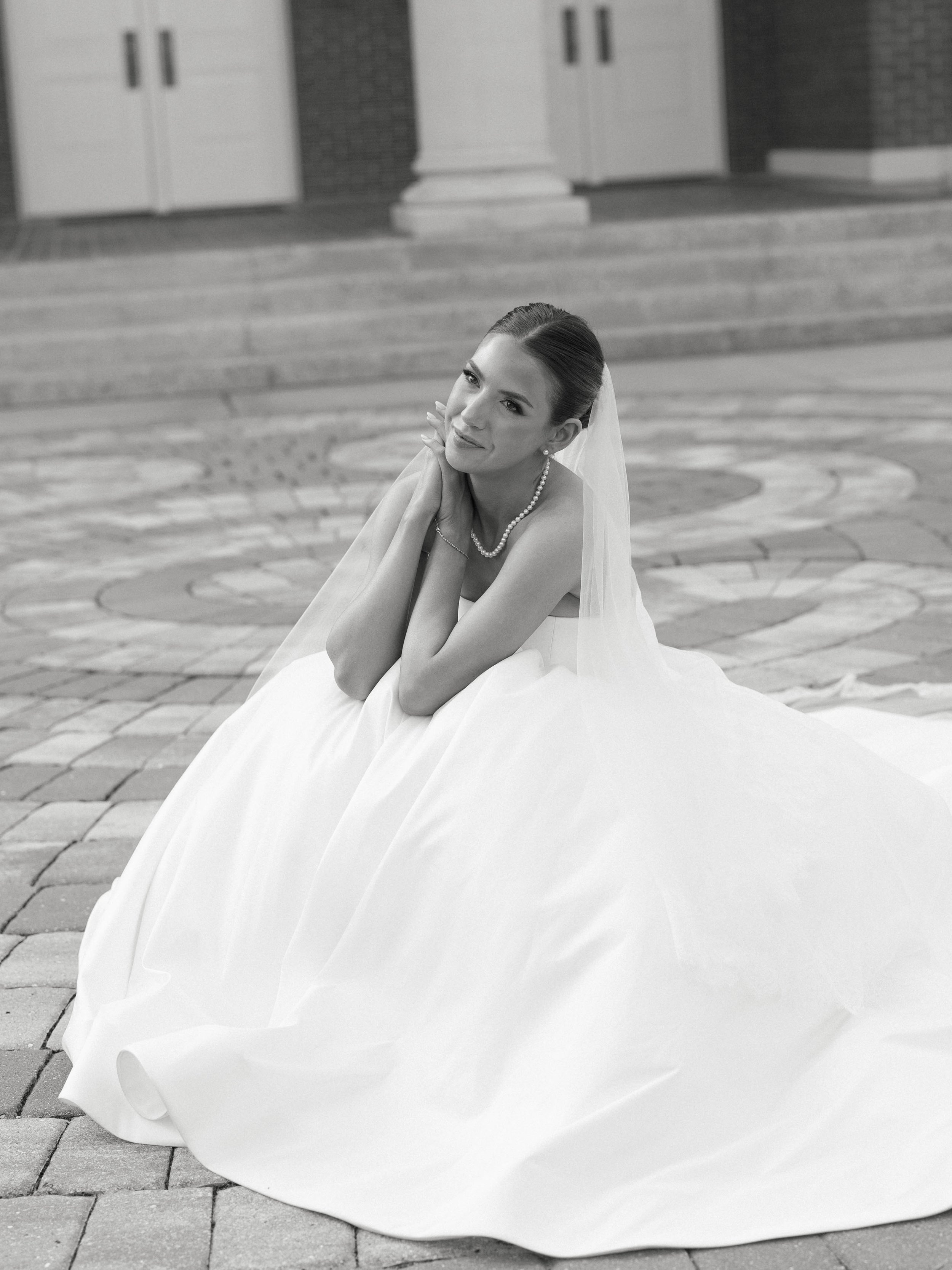 A bride sitting outdoors on a paved surface, dressed in a wedding gown with a veil and pearl necklace, smiling softly at the camera.