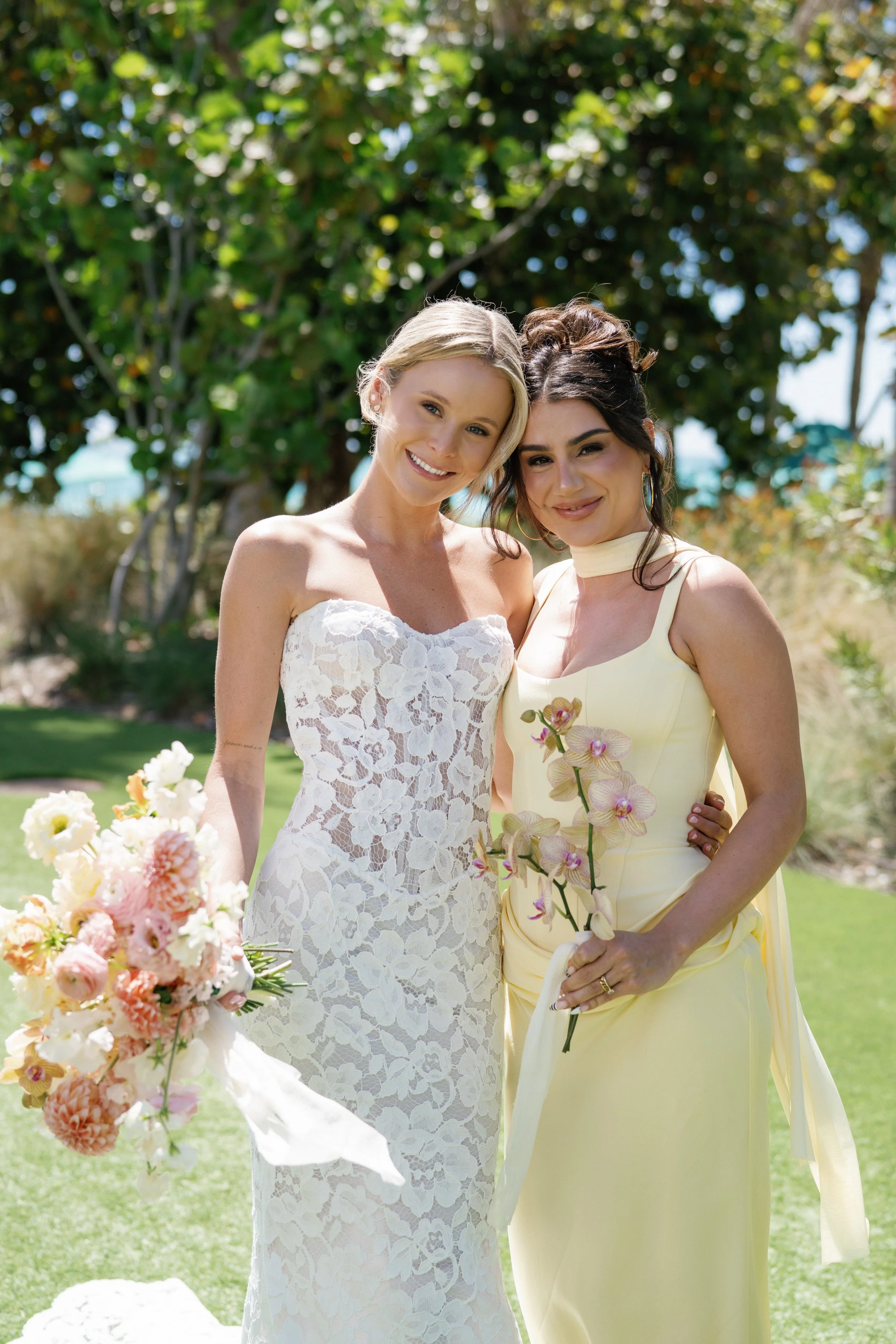 Two women, one in a white lace wedding dress and the other in a yellow dress with floral accents, smiling outdoors with trees and flowers in the background.