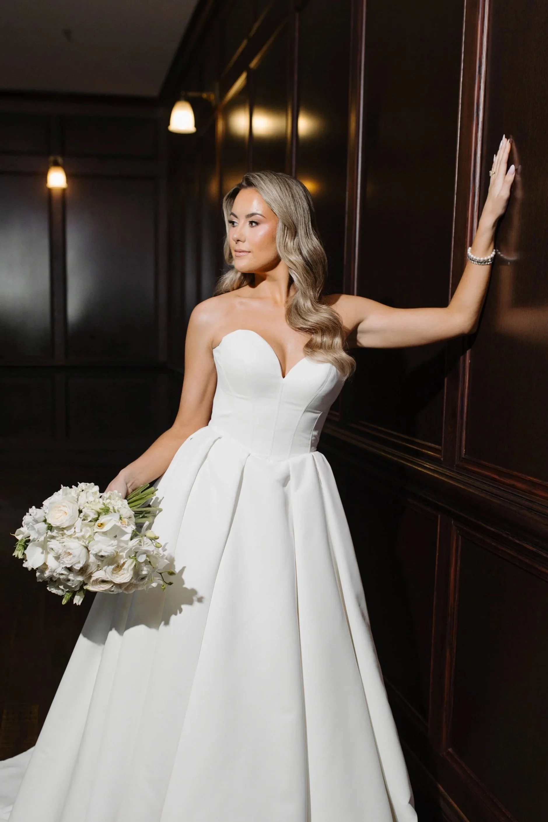 A woman in a white wedding dress holding a bouquet of white flowers, leaning against a dark wooden wall with three hanging lights in the background.