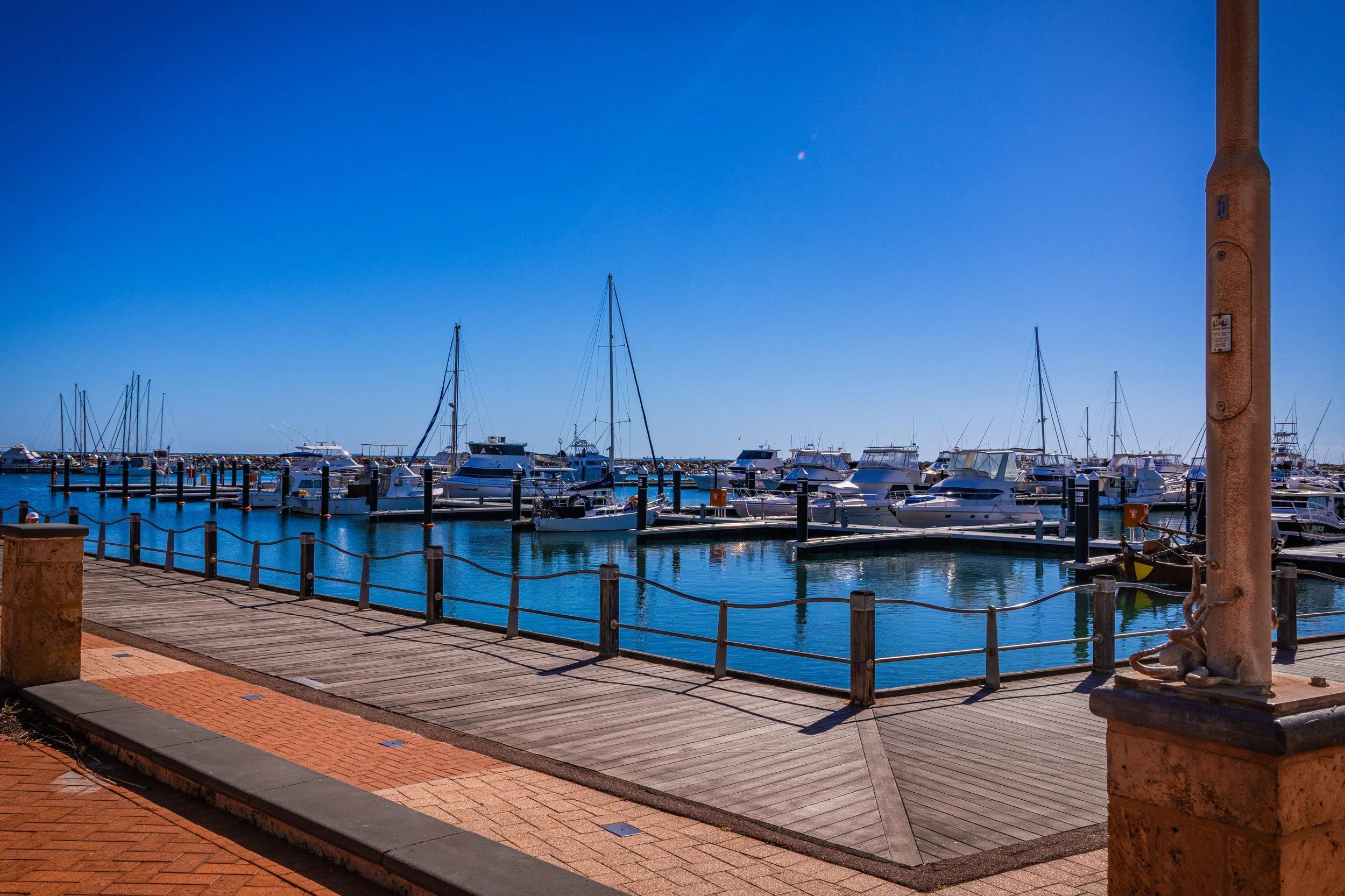 Geraldton marina with boats docked at the pier under clear blue sky, viewed from a walking path with brick and wooden pavement, and a post with an octopus on the right side. Photographer Elsa Norvill