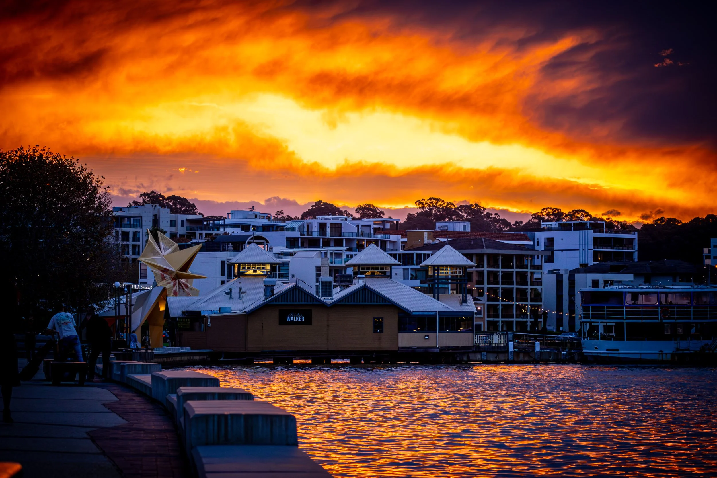 Sunset over the waterfront at South Perth, looking towards Kings Park in Western Australia, with modern white buildings, dockside benches, a boat, trees, and a bright orange and yellow sky with dark clouds. Photographer Elsa Norvill