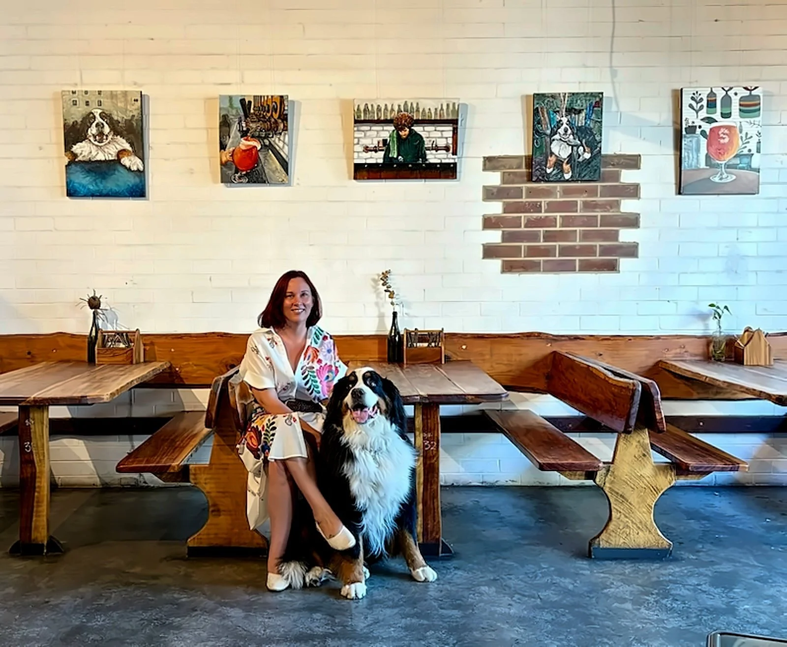 A woman with red hair wearing a white dress with colorful floral patterns sitting at a wooden table with a large black and white Bernese Mountain Dog in a cozy cafe with white brick walls and colorful artwork hanging above.