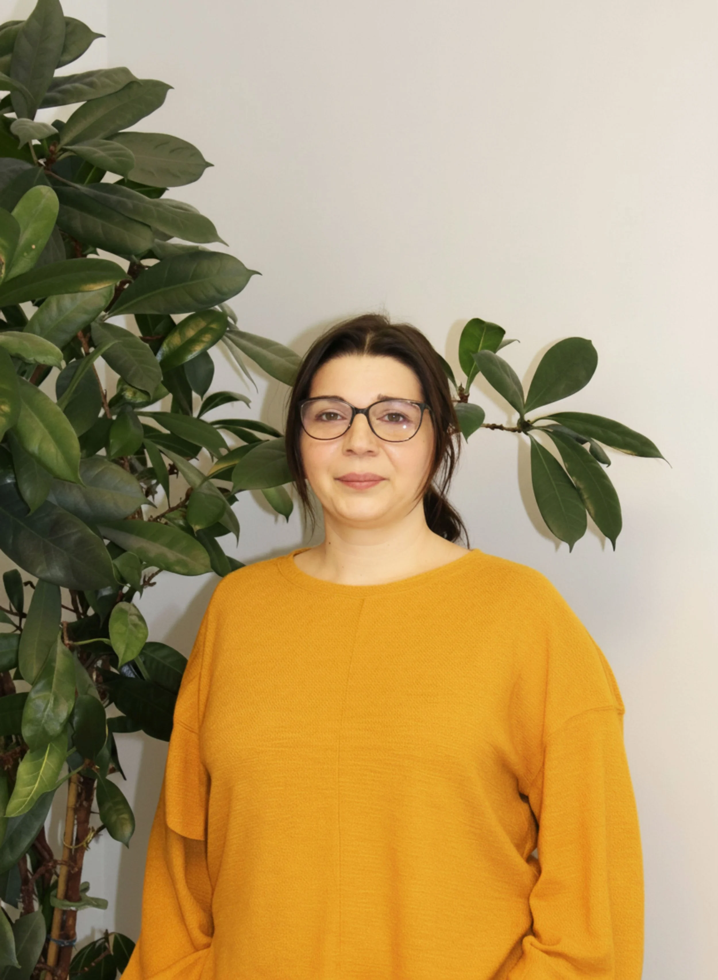 Eine Frau mit braunen Haaren, Brille und einem orangefarbenen Pullover, steht vor einer weißen Wand und einem grünen Zimmerpflanzen.
