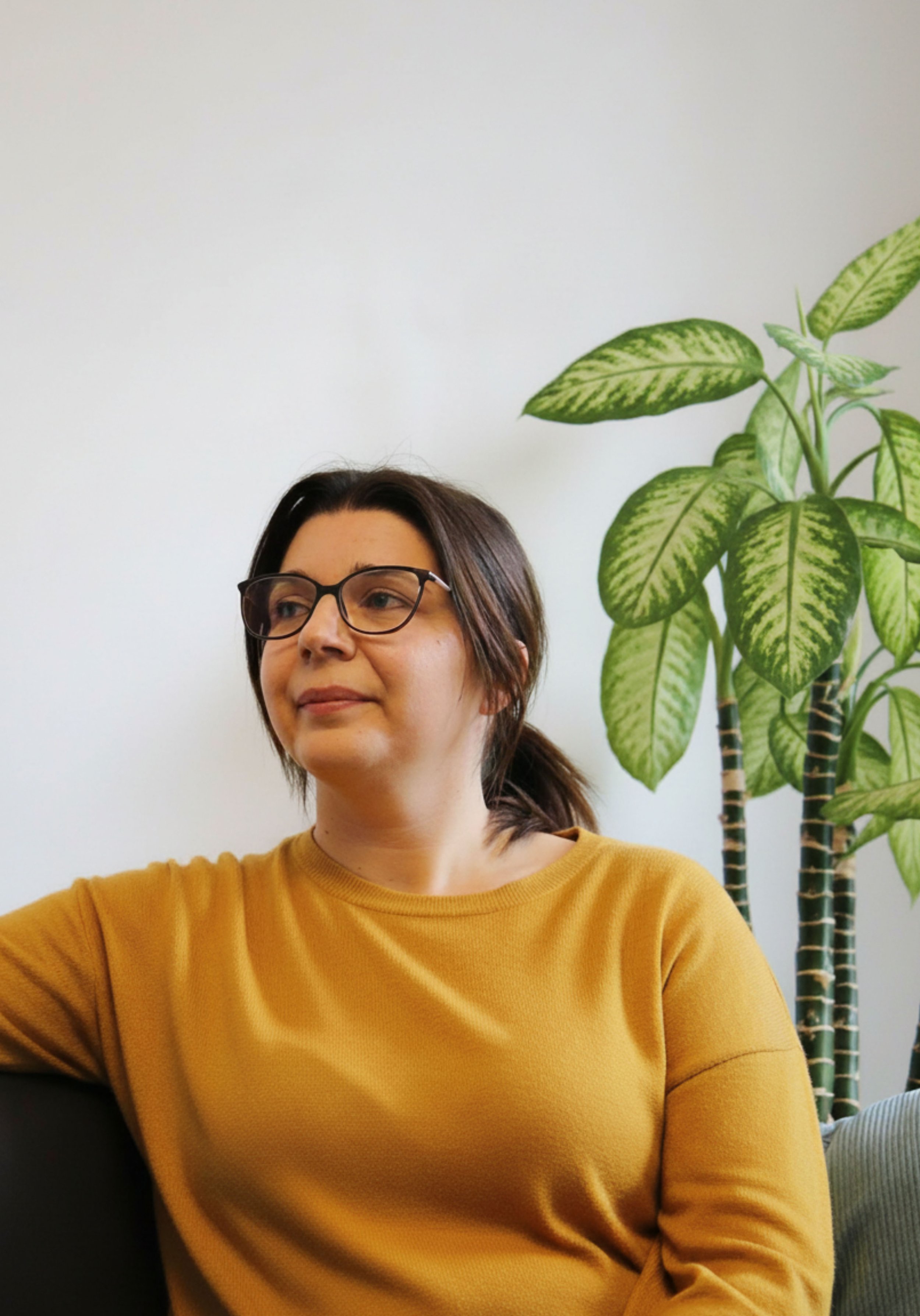 Eine Frau mit braunen Haaren, Brille und einem senffarbenen Pullover sitzt vor einer weißen Wand mit einer großen grünen Zimmerpflanze.