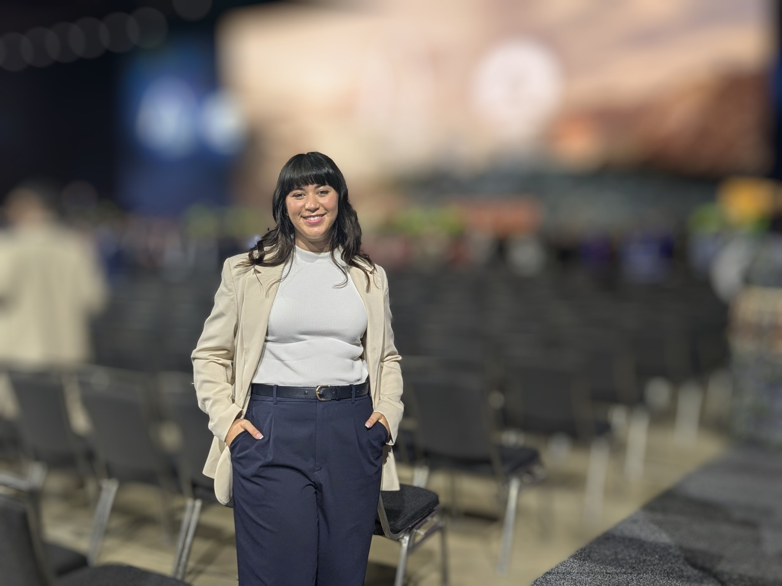 A woman with dark hair and bangs, smiling, standing in an airport or train station, with blurred background of chairs and lights.