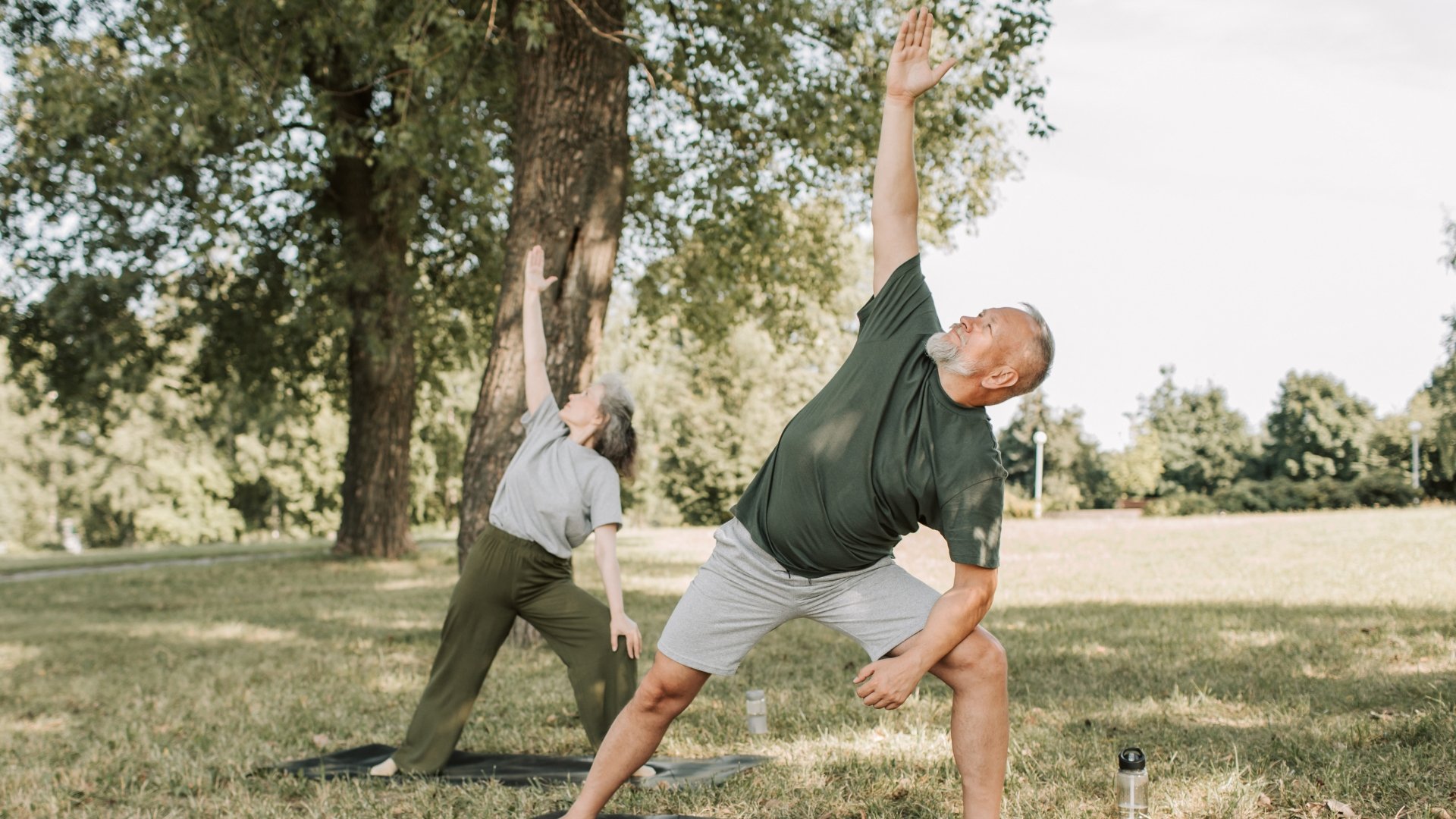 An elderly man and woman practicing yoga outdoors in a park during daytime. They are both in a wide stance pose, reaching one arm upward. The man is wearing a dark green shirt and gray shorts, the woman is wearing a gray shirt and green pants. The background features trees with green leaves, and the ground is grassy. There are water bottles on the ground nearby.