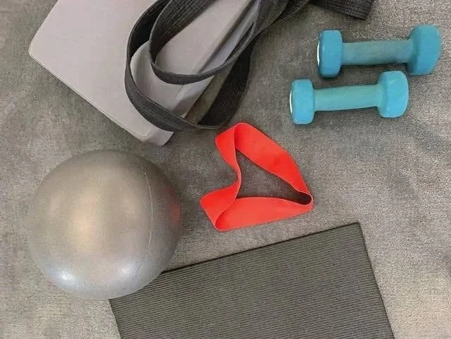 Exercise equipment including two light blue dumbbells, a red resistance band, a silver exercise ball, a black and white yoga mat, and a gray foam roller on a gray carpeted floor.