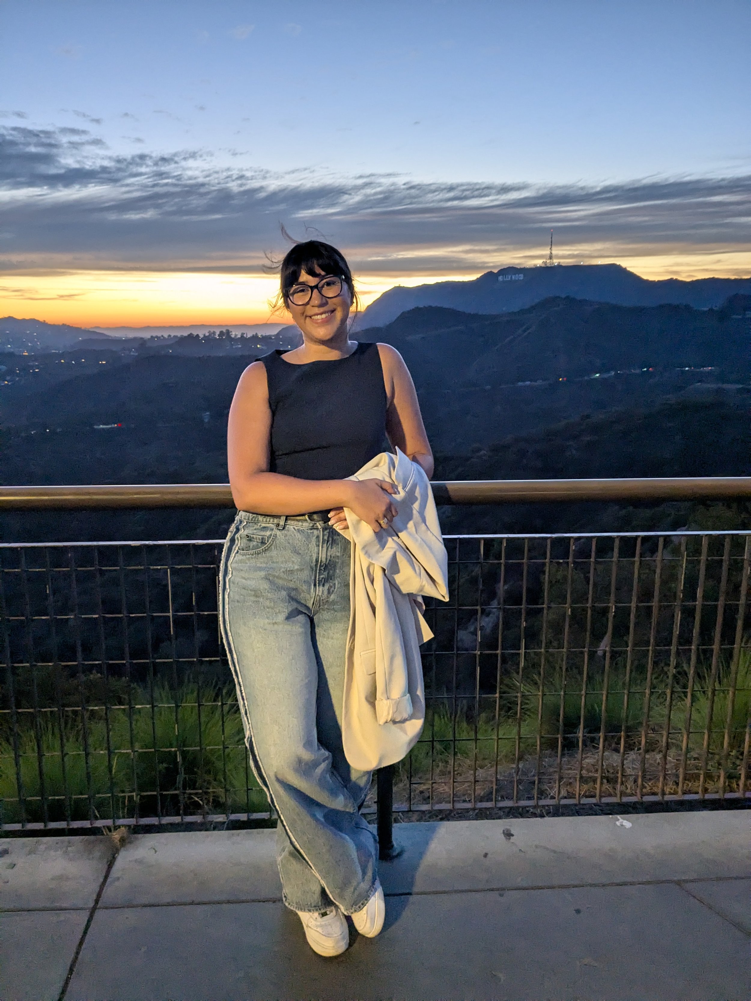 A woman wearing glasses, a black sleeveless top, and jeans, smiling while holding a beige jacket, standing in front of a sunset view of the Hollywood sign on a hill, with a metal railing in the foreground.