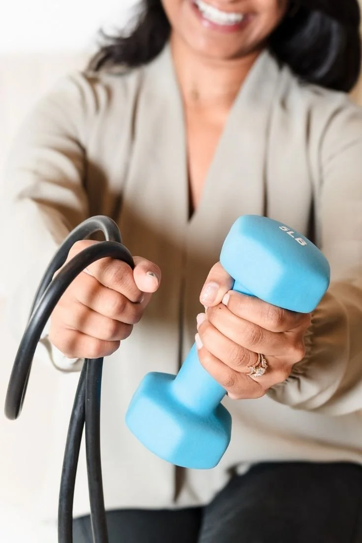 A woman smiling and holding a yellow dumbbell in one hand and a black jump rope in the other.