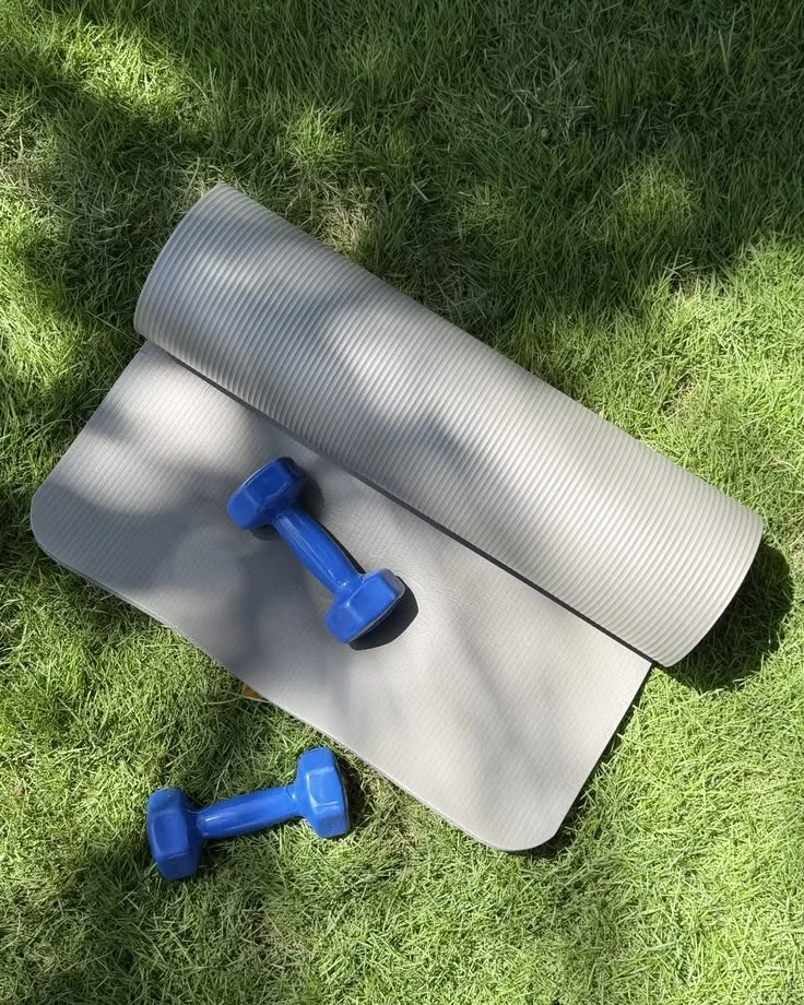 Gray yoga mat partially rolled out on green grass with two small blue dumbbells placed on it, one on the mat and one beside it.