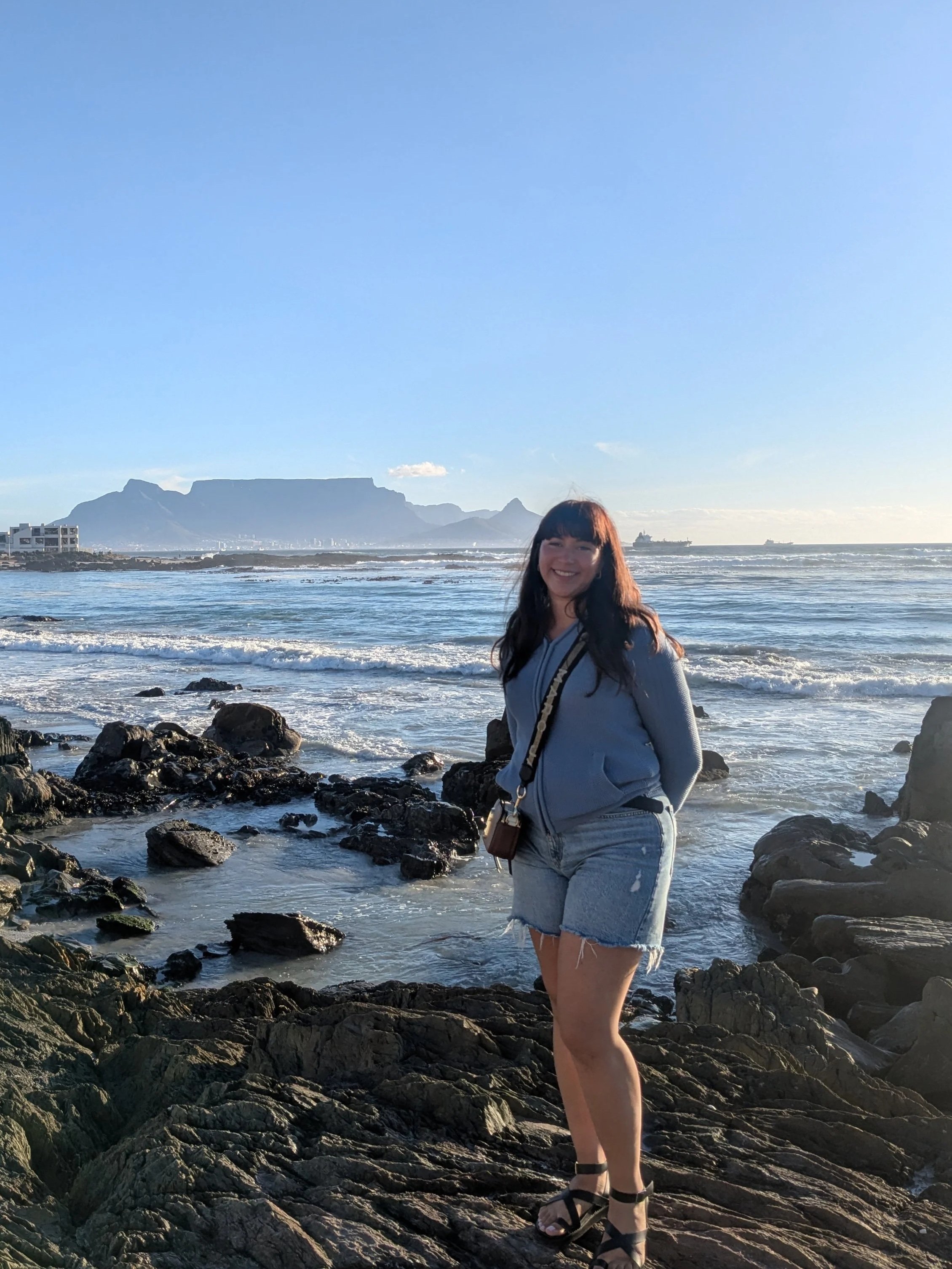 A woman standing on rocks at the beach with the ocean and mountains in the background, smiling and wearing a gray hoodie, denim shorts, and sandals.