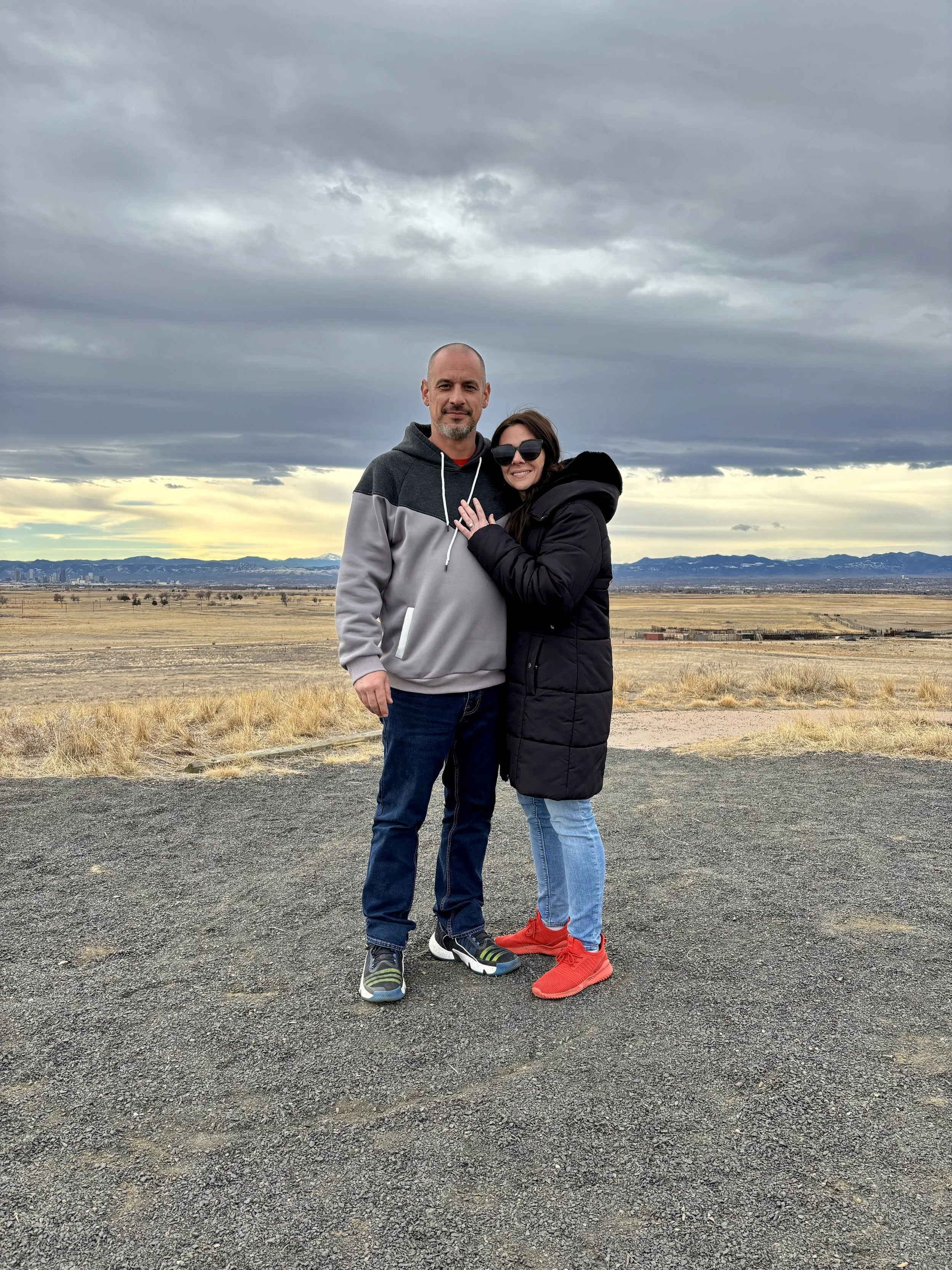 A man and woman standing close together outdoors on a paved area with a vast open landscape, mountains, and cloudy sky in the background. The woman is wearing sunglasses, a black puffy jacket, light blue jeans, and red sneakers, while the man is wearing a gray and black hoodie, blue jeans, and multicolored sneakers. They are smiling and appear happy.