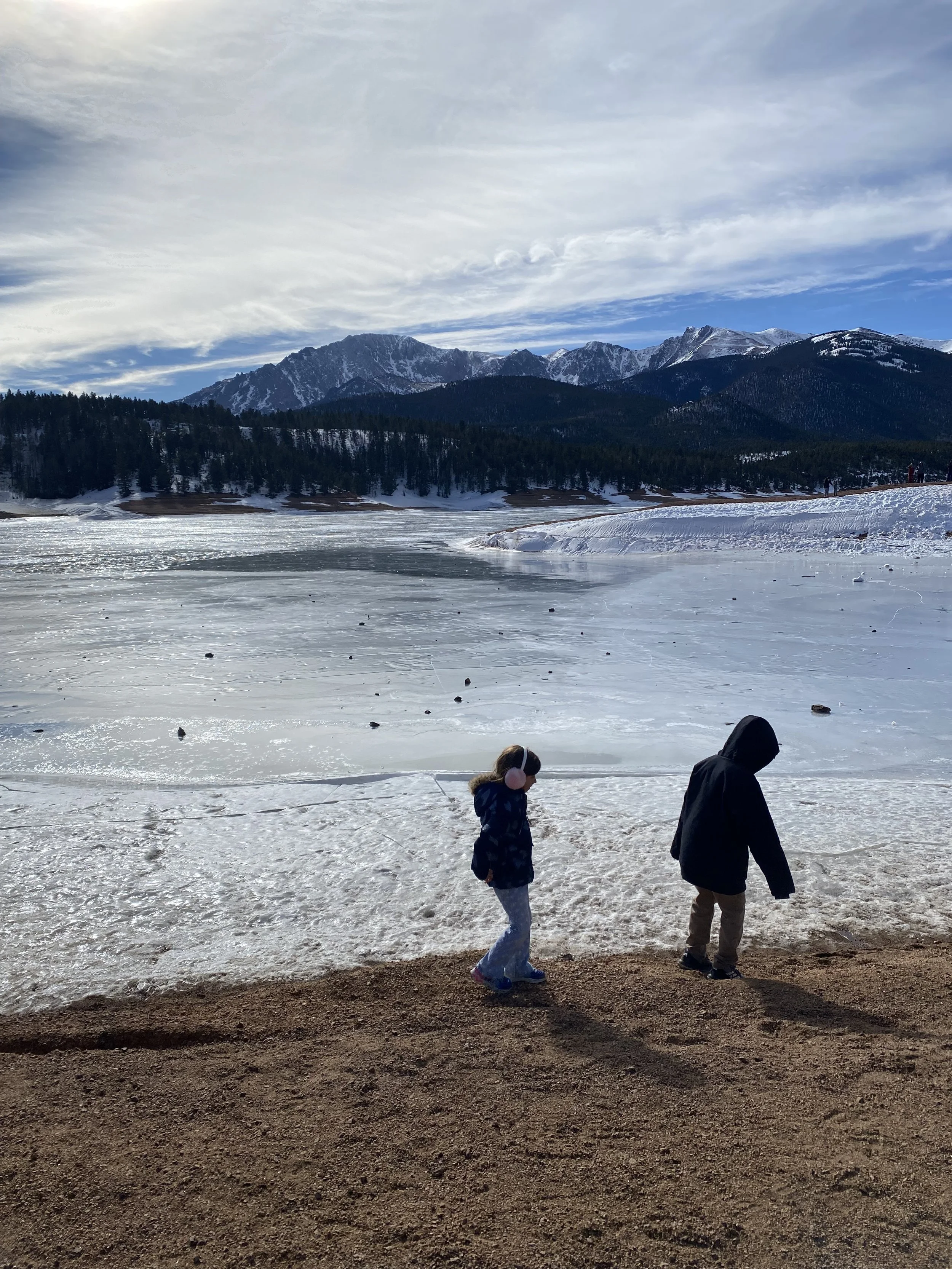 Two children walking along a snowy shore with a frozen lake, forested hills, and snow-capped mountains in the background under a partly cloudy sky.