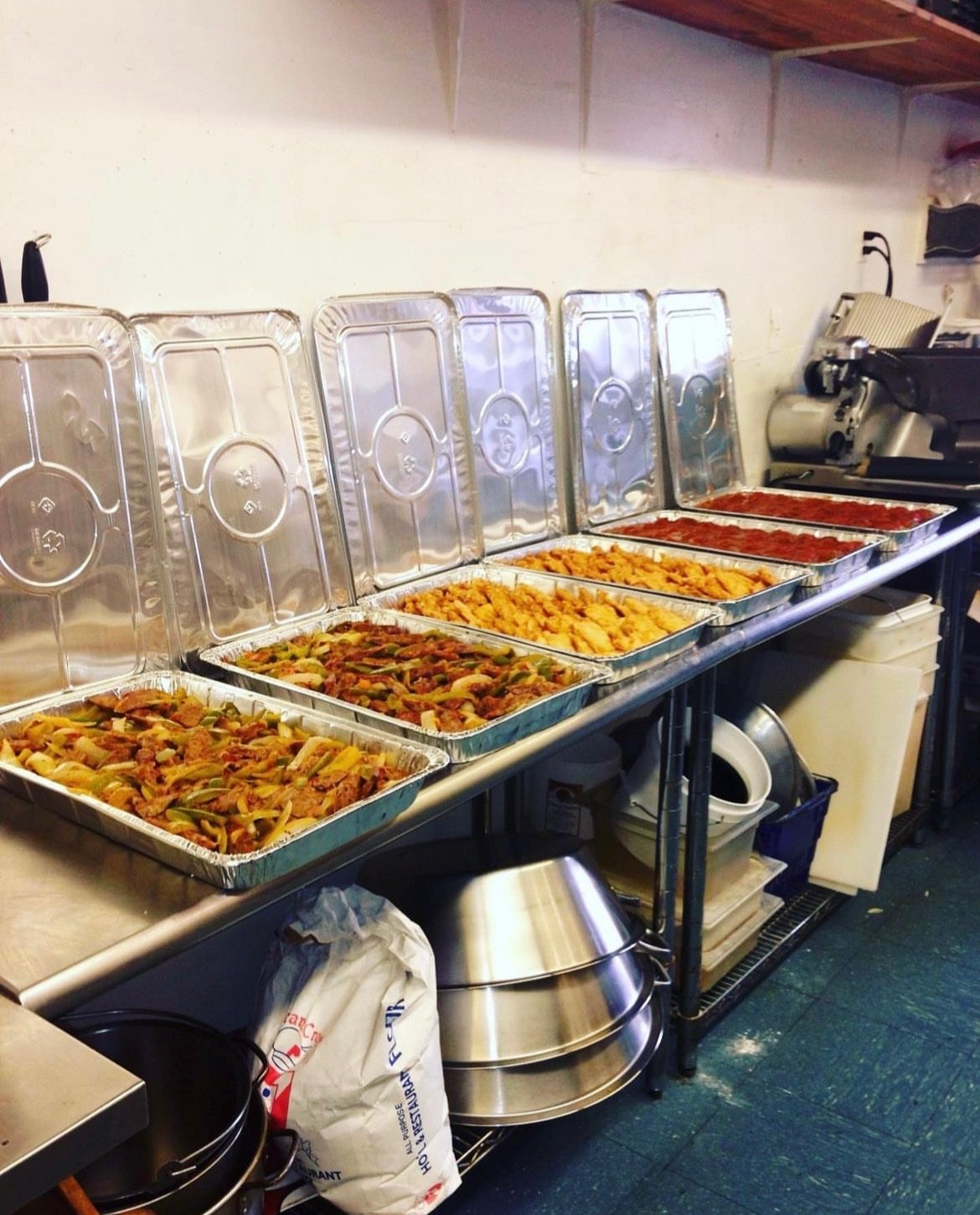 Six aluminum trays of cooked food with clear plastic lids on a counter, including vegetable stir-fry, fried chicken, and two trays of tomato sauce, with kitchen appliances and storage underneath.