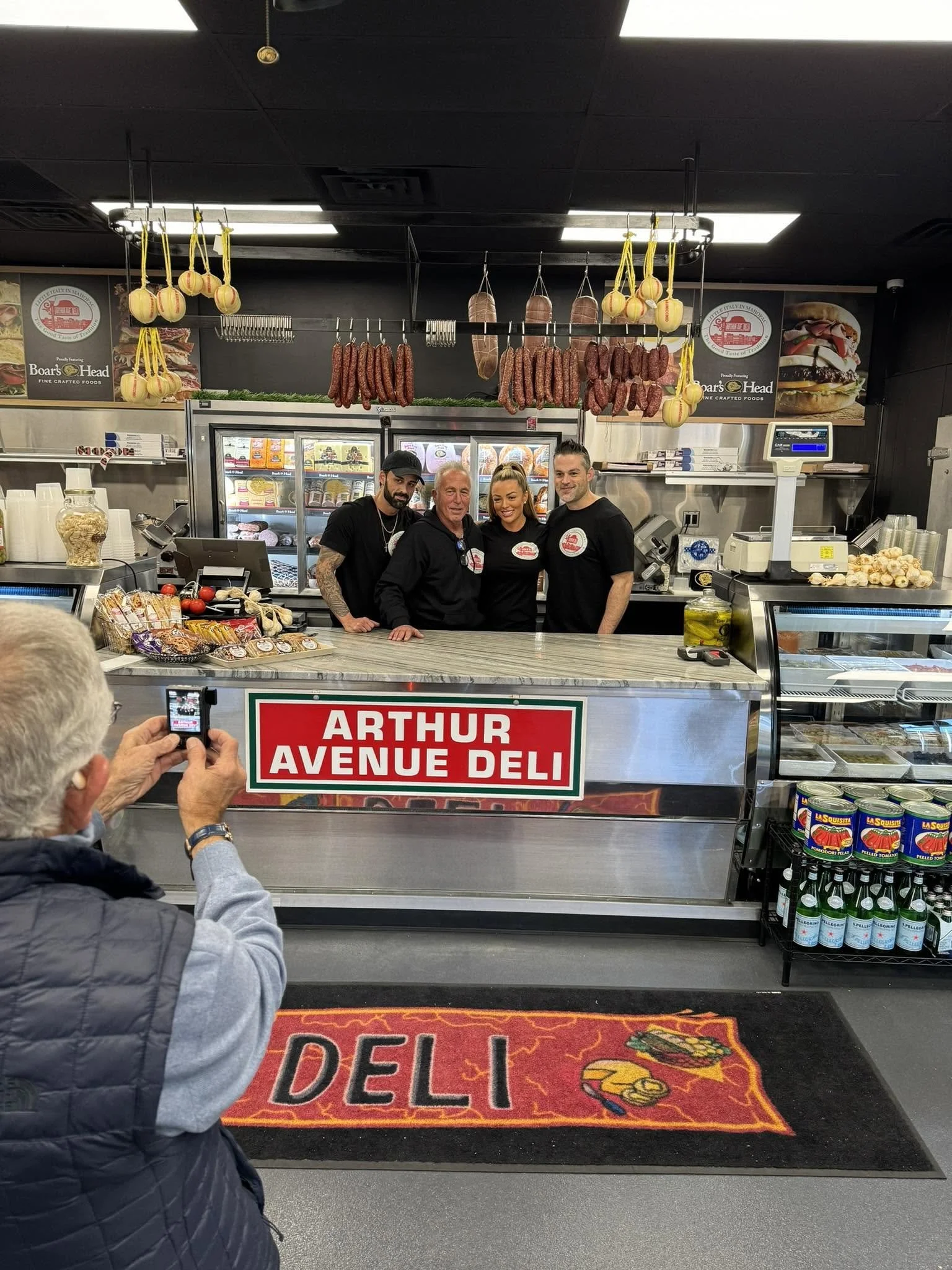 Four people standing behind the counter at Arthur Avenue Deli, with hanging sausages and meats above them. A person in the foreground is taking a photo with a phone. There are food items and condiments on the counter and shelves behind them.