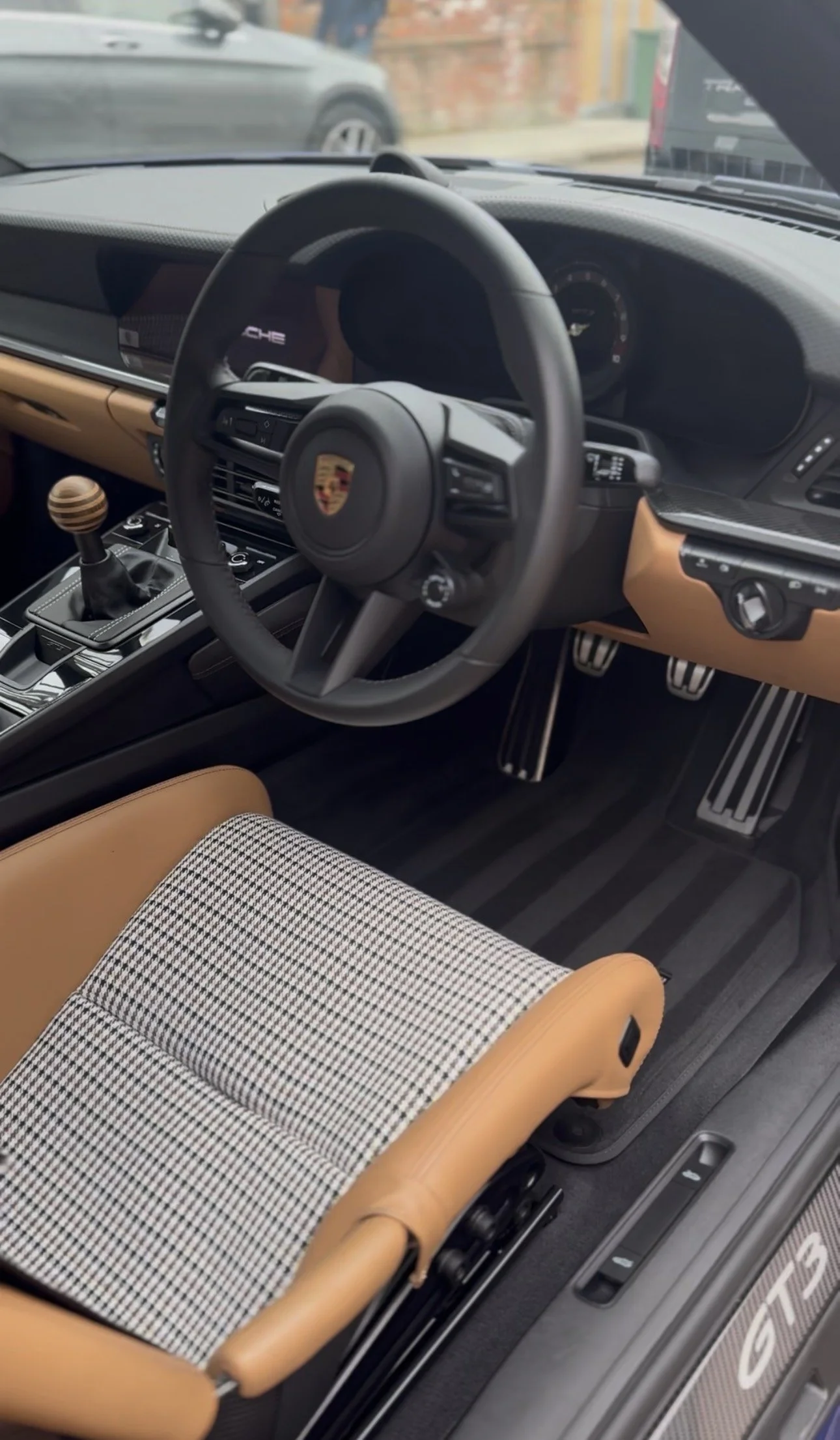 Interior of a Porsche sports car showing the steering wheel, dashboard, gear shifter, and front seat with a plaid pattern and tan leather accents.