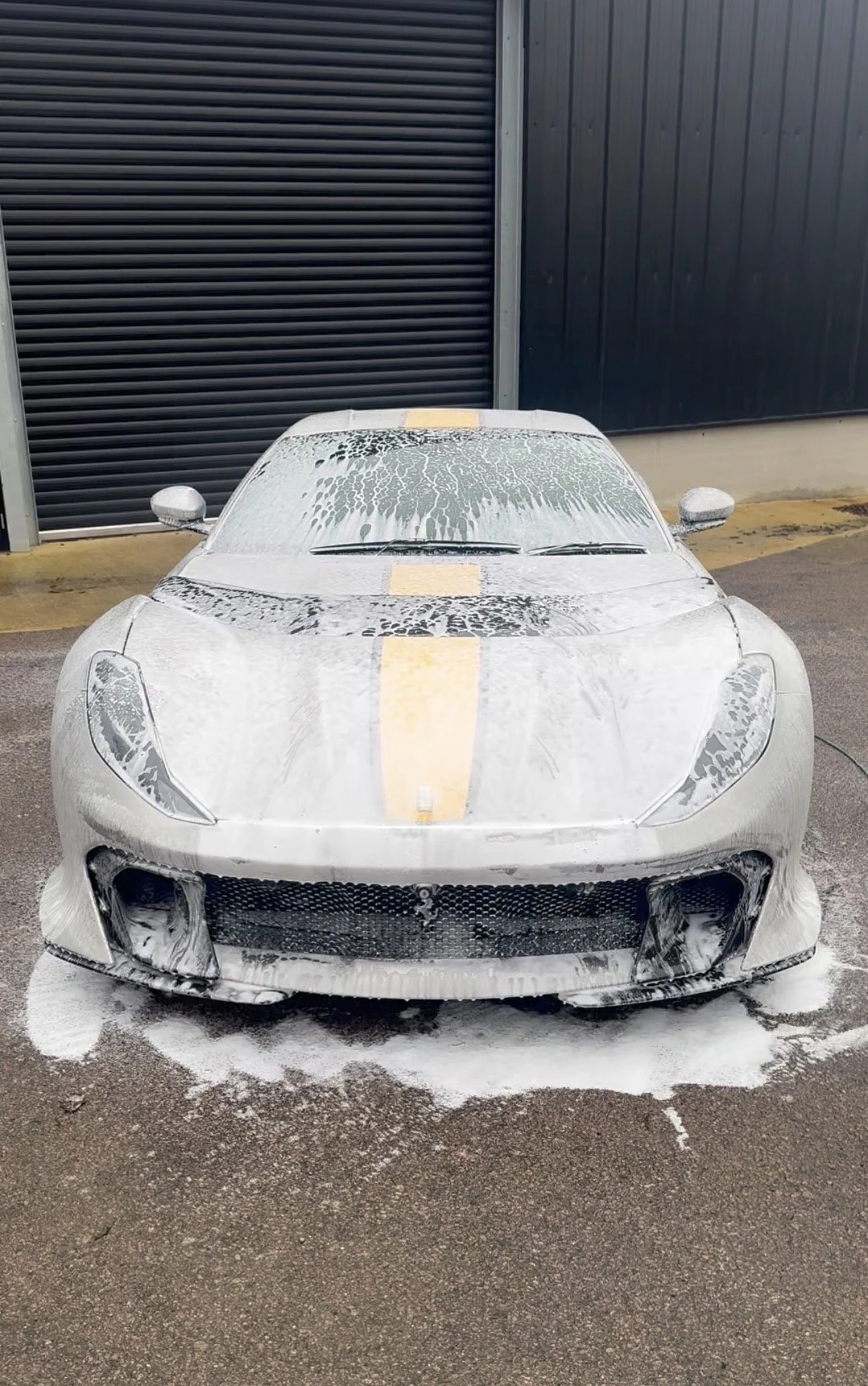 White sports car covered in soap suds during a car wash, parked in front of a black metal building.