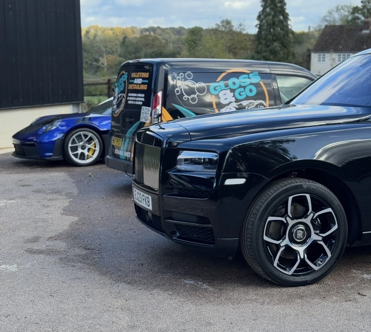 Several luxury cars parked side by side outdoors, including a black Rolls-Royce with distinctive wheels, a blue sports car, and a black utility van with colorful advertising for a vehicle detailing service.