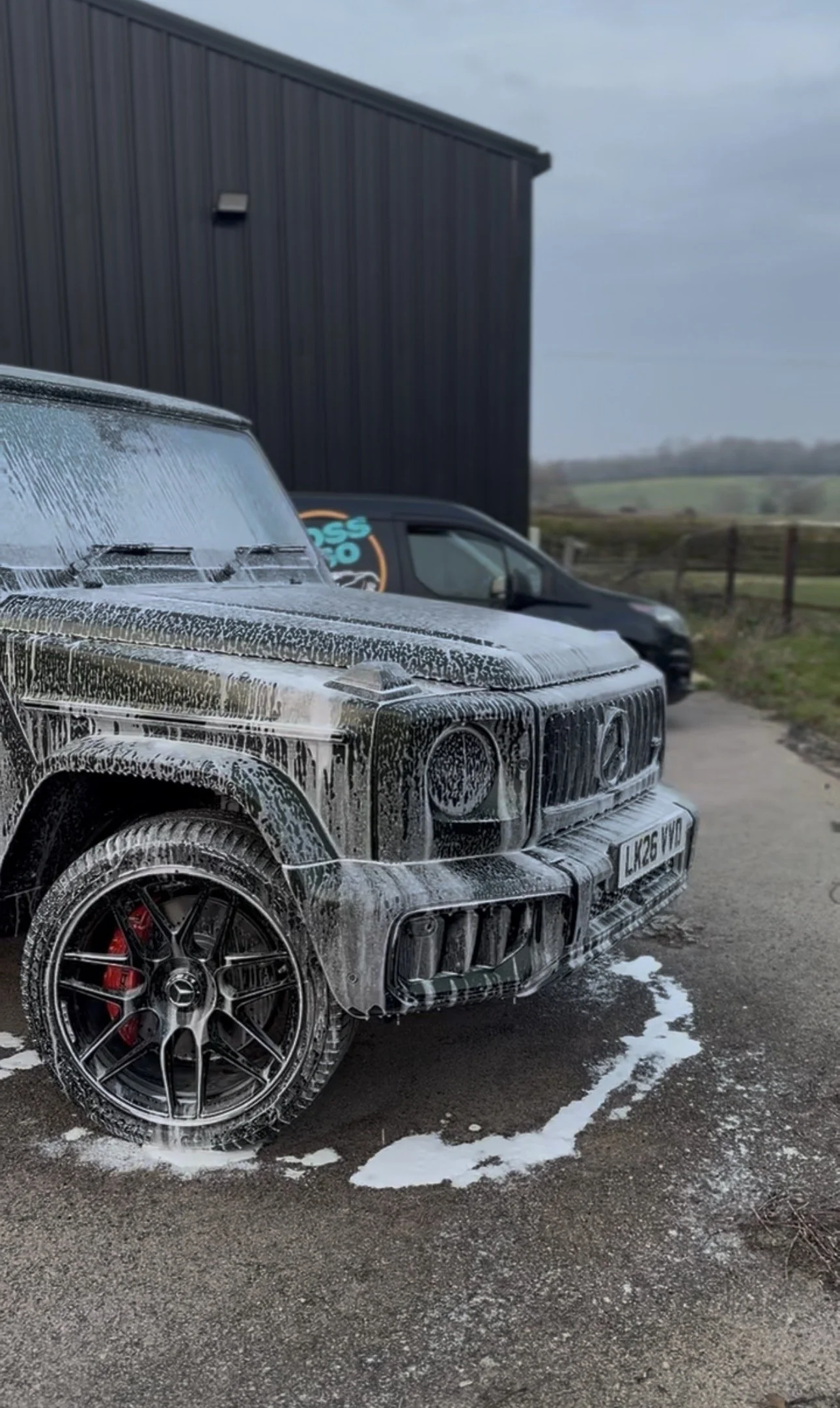 A black luxury SUV covered in soap suds during a car wash, parked outdoors near a dark building with a rural landscape in the background.
