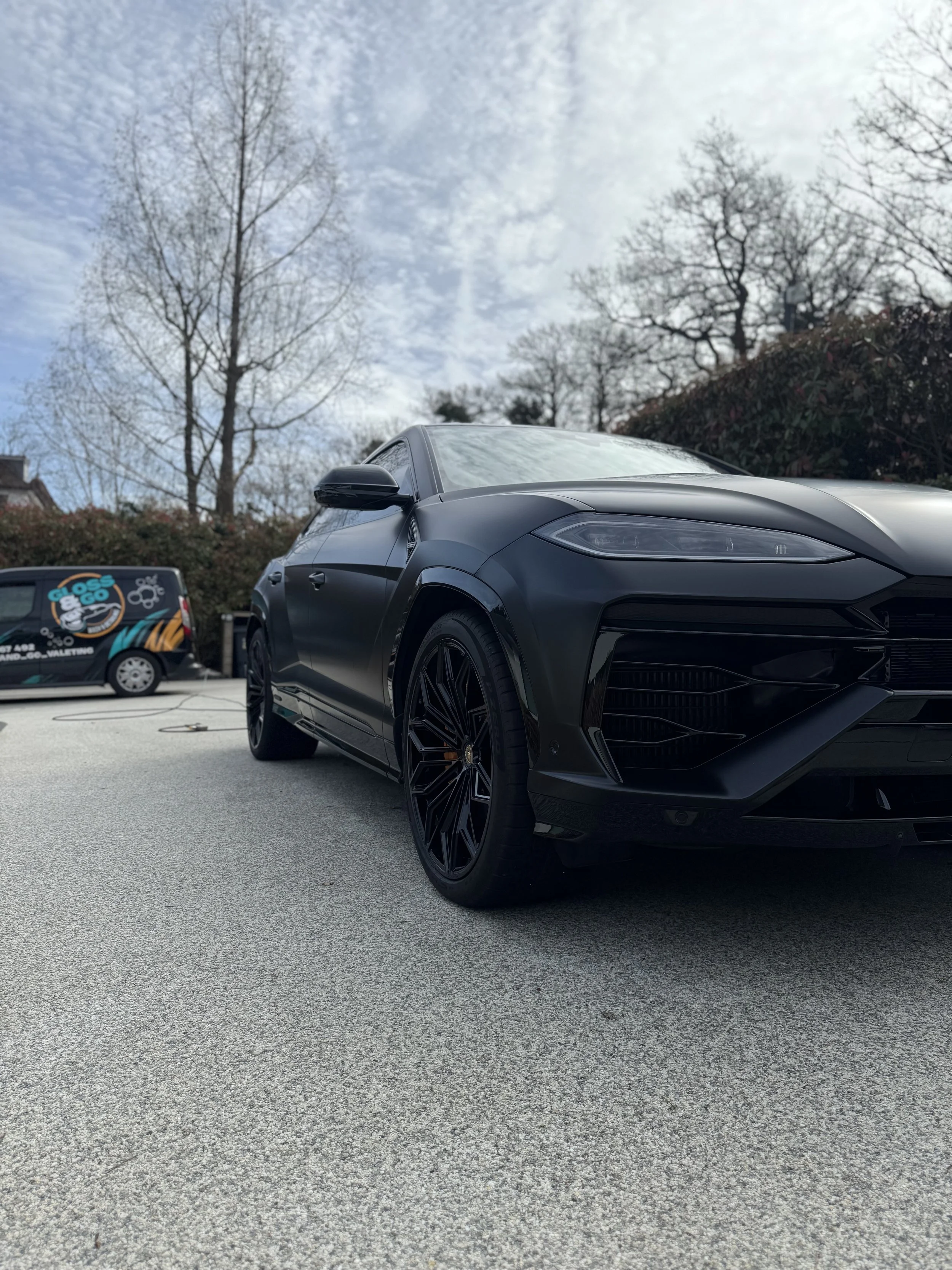 A black luxury car parked in an outdoor parking lot with trees and a cloudy sky in the background.