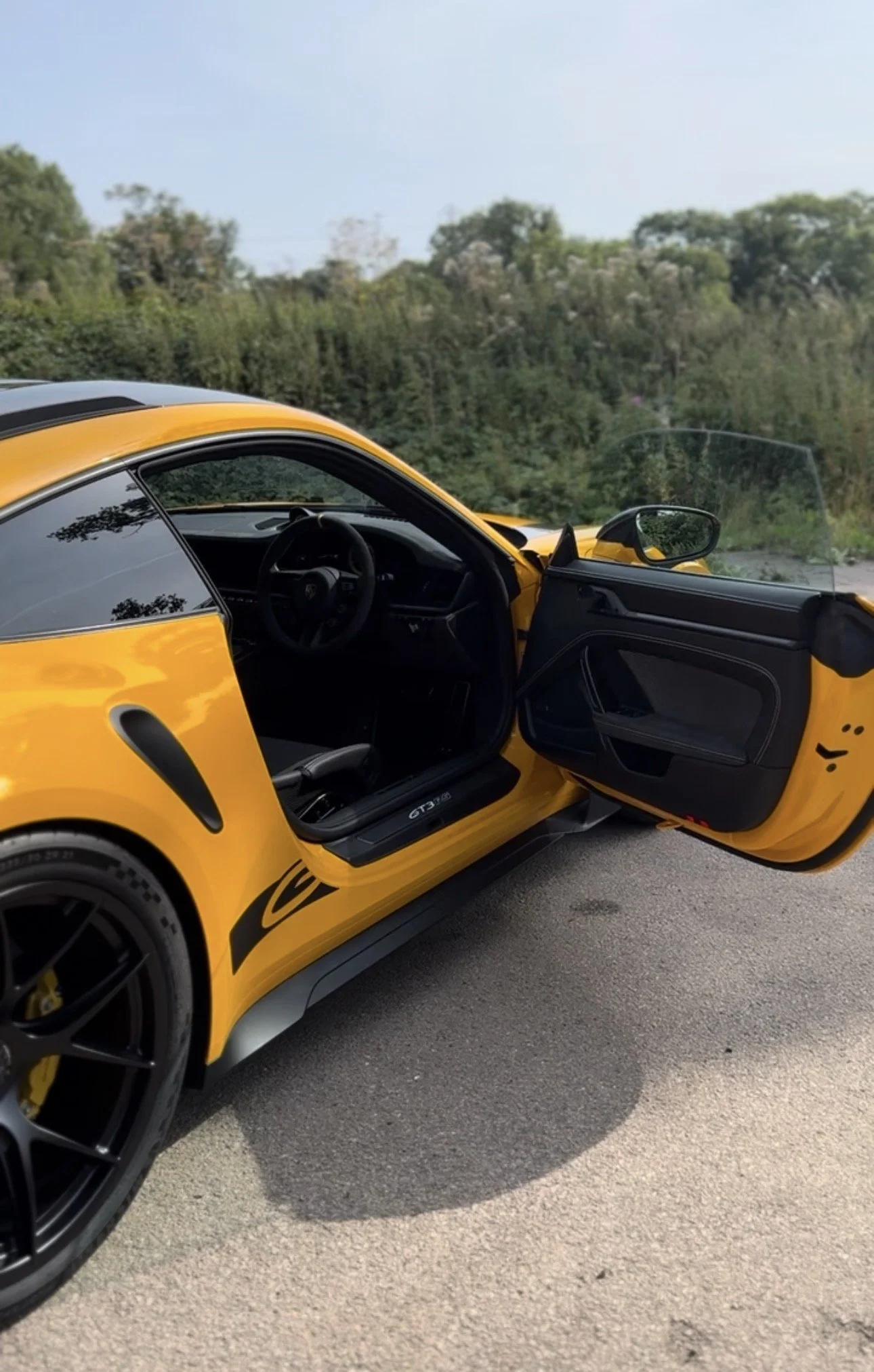 A yellow sports car with black wheels and a black interior, parked on a road with an open door. The background features trees and a blue sky.