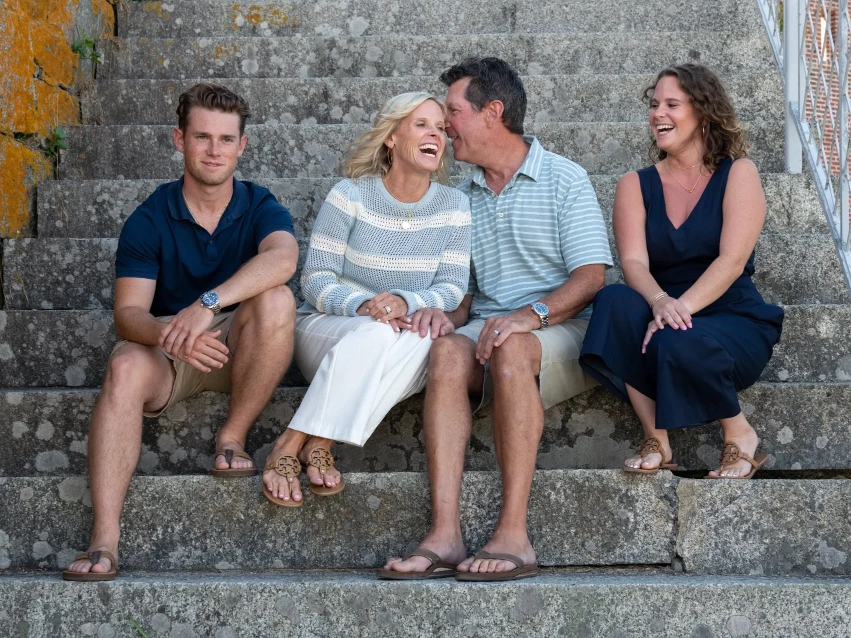 Four people sitting on outdoor stone stairs, engaged in conversation and laughter. The group includes two young women and an older woman and man, dressed casually in summer clothing.